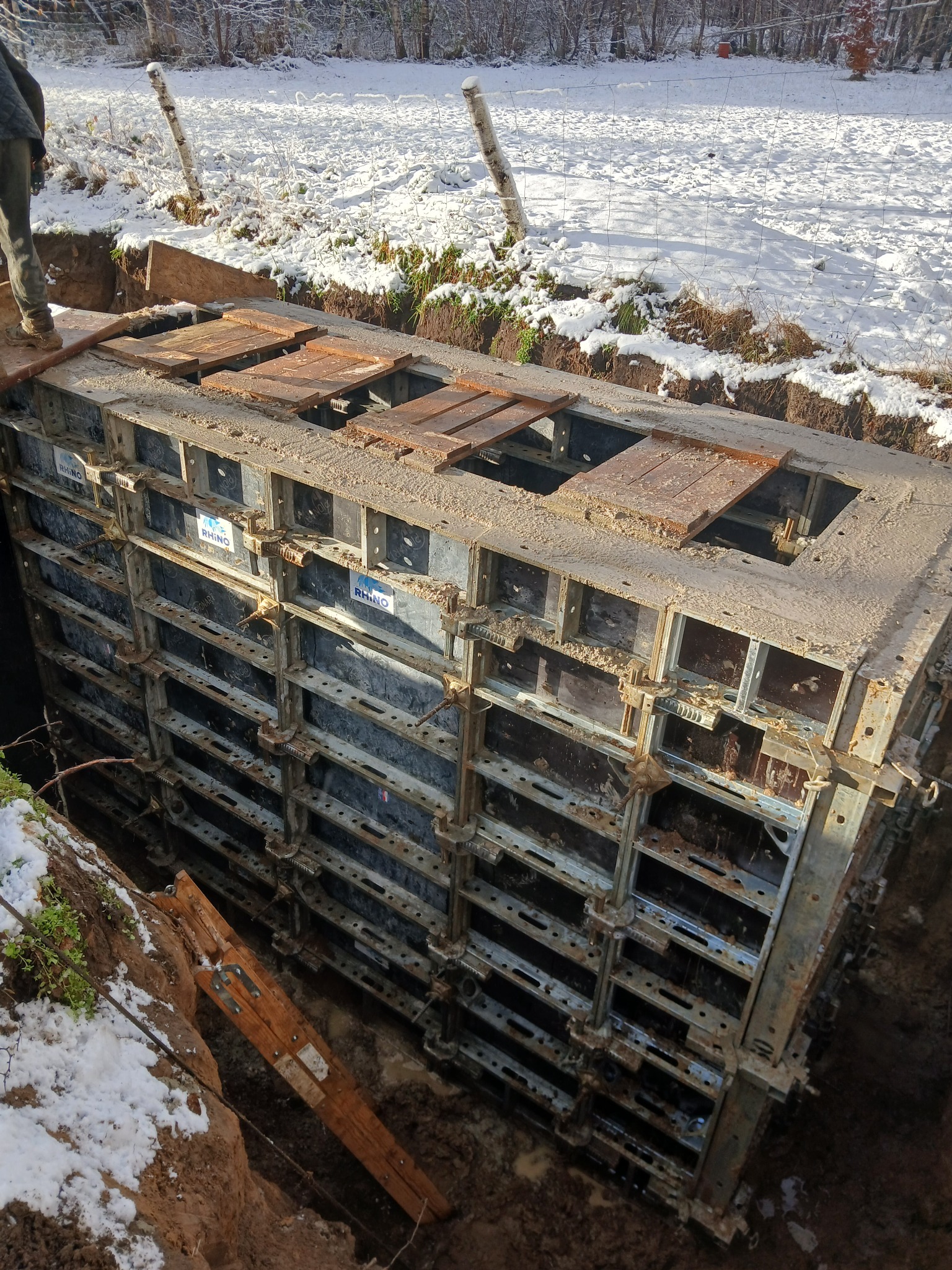 Szare szalunki fundamentowe w wykopie, częściowo przykryte drewnianymi deskami. W tle ziemia pokryta śniegiem i drzewa.