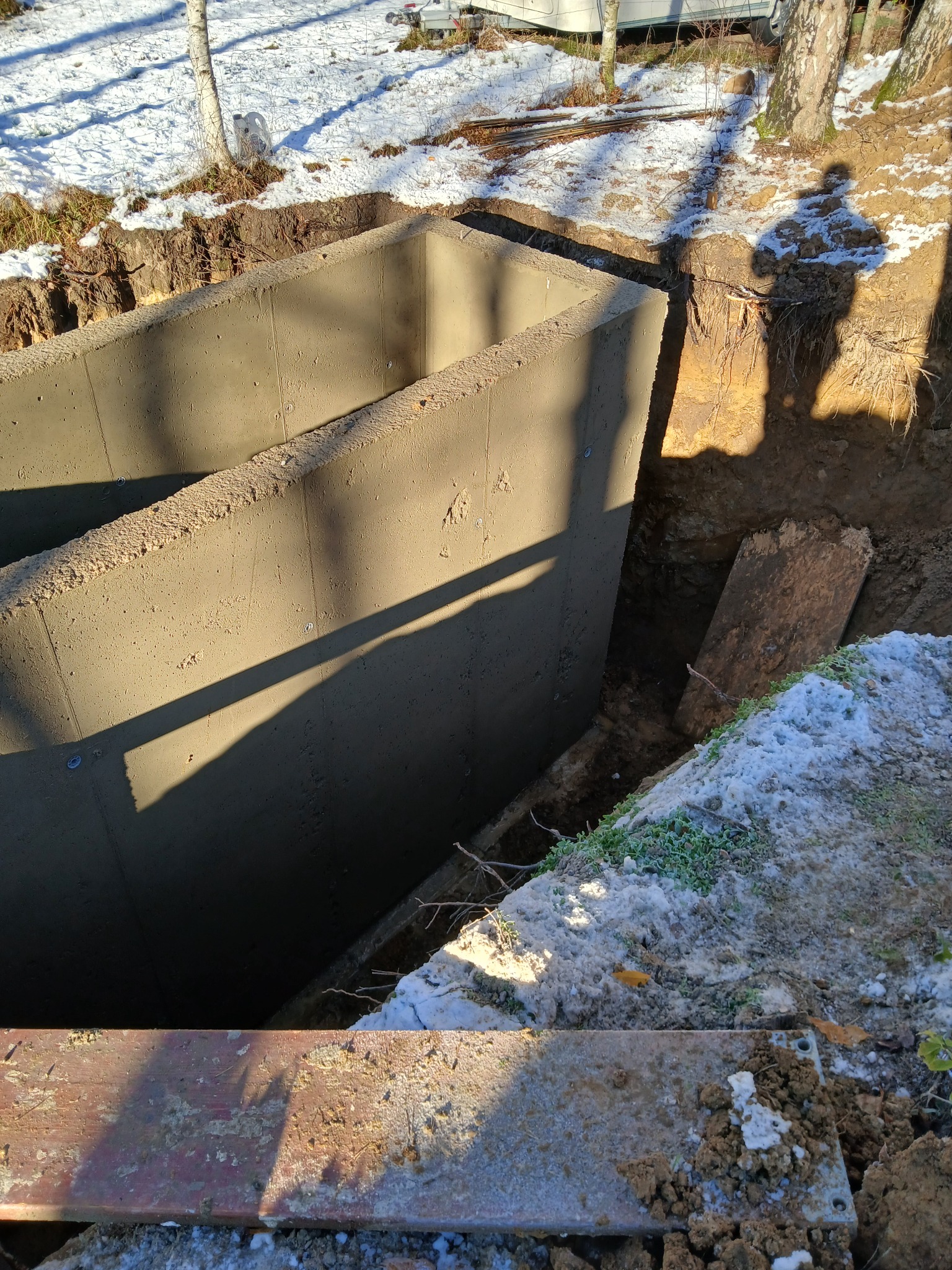 Surowy betonowy fundament w wykopie, widok z góry, częściowo pokryty śniegiem. W tle drzewa i cień osoby.