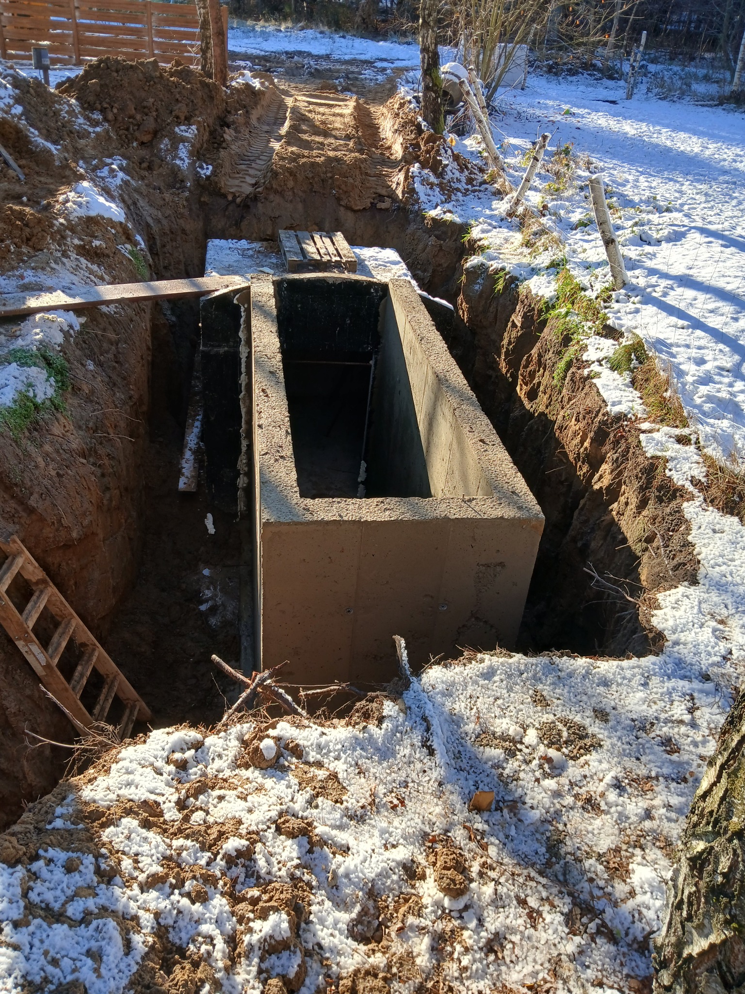Betonowa konstrukcja w wykopie ziemnym, częściowo zasypana śniegiem. Widoczne ślady opon i drewniana drabina. Prace ziemne w zimowej scenerii.