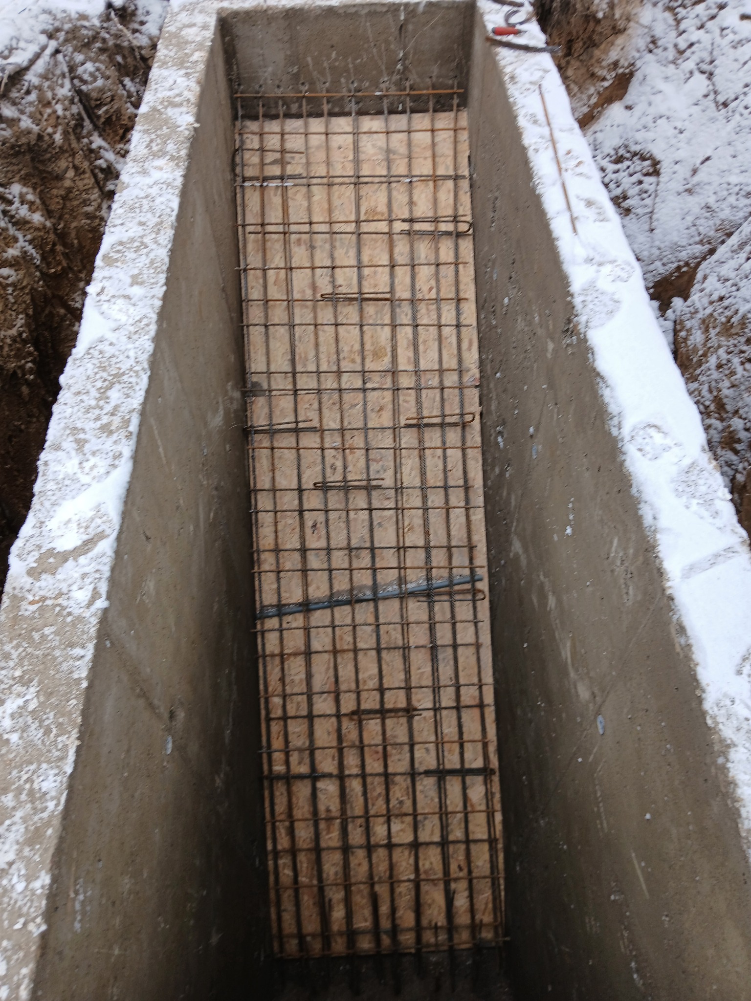 Zbrojenie w szalunku fundamentu, widok z góry. Świeży beton i śnieg na krawędzi wykopu. Siatka zbrojeniowa na drewnianym dnie. Etap budowy fundamentów.