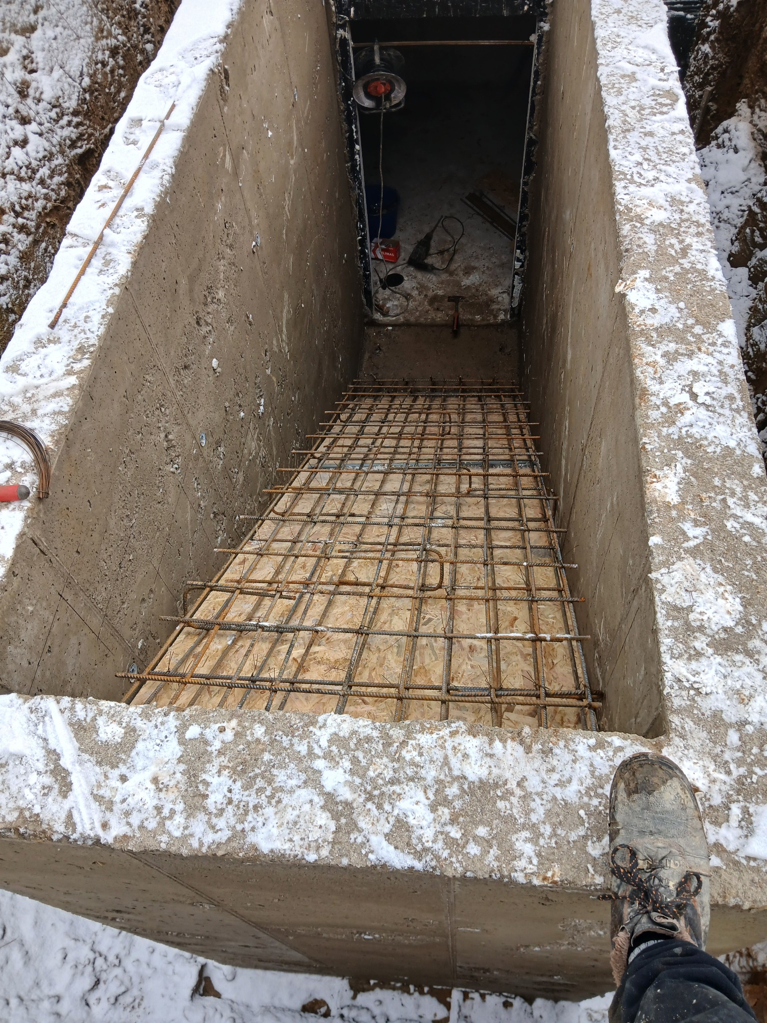 Zbrojenie w betonowym wykopie fundamentowym, częściowo pokrytym śniegiem. Widoczne narzędzia i fragment buta pracownika. Ujęcie z góry.