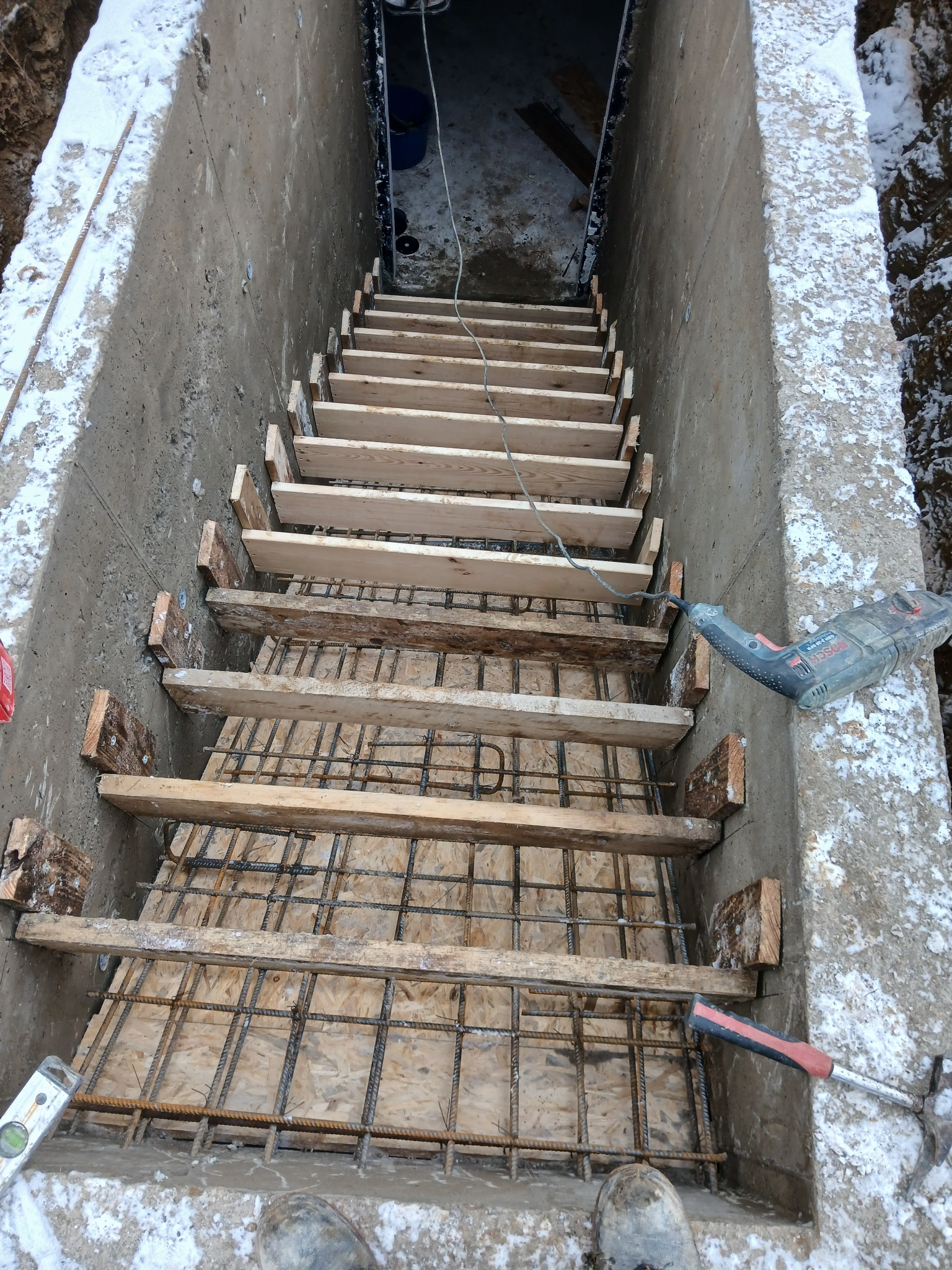 Zbrojenie schodów w wykopie fundamentowym, widok z góry. Stalowe pręty, drewniane szalunki i narzędzia budowlane w zimowym otoczeniu. Etap prac fundamentowych.