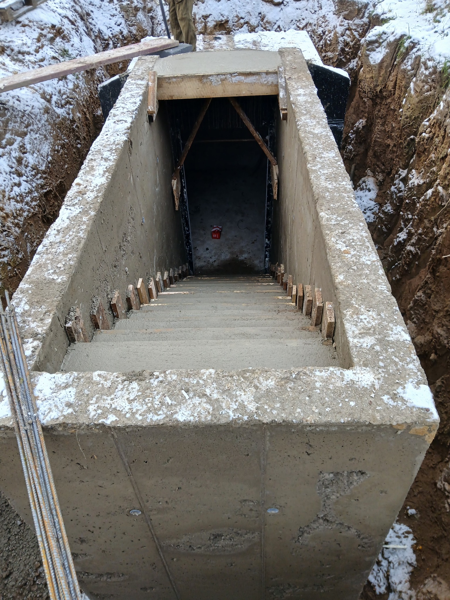 Betonowa konstrukcja schodów w wykopie, widok z góry. Ślady śniegu na betonie, drewniane elementy szalunku. W tle ziemia i fragment ubrania osoby.