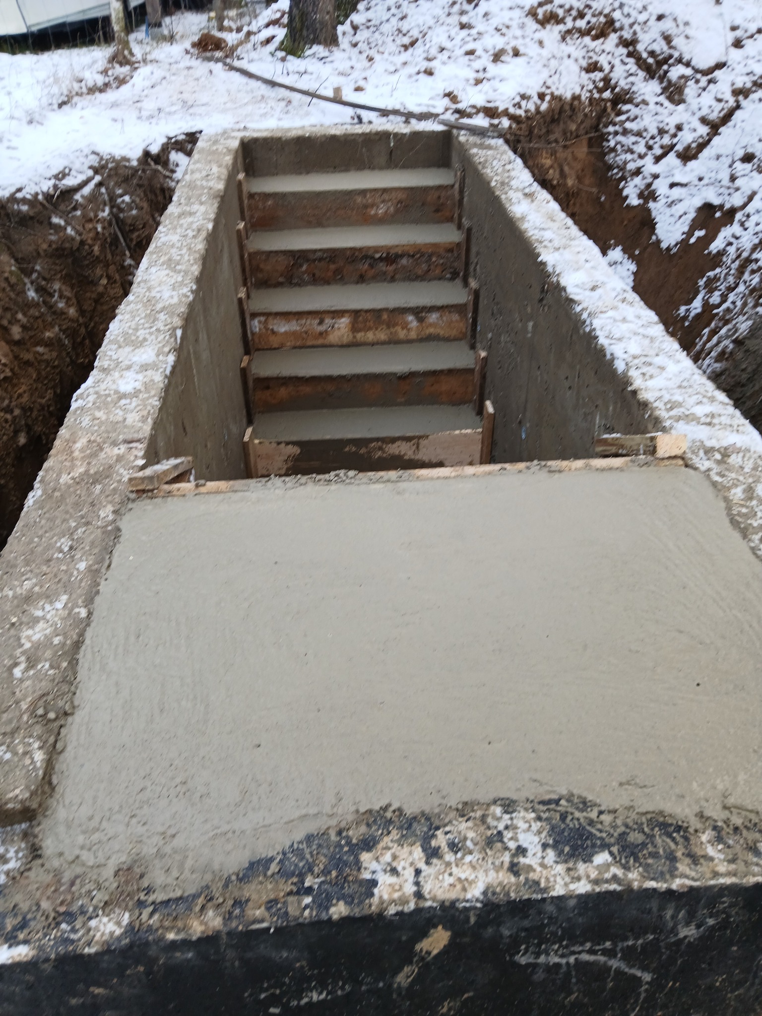 Betonowa konstrukcja schodów w wykopie, świeżo wylana, z widocznymi drewnianymi elementami szalunku. Zimowy krajobraz z resztkami śniegu w tle.