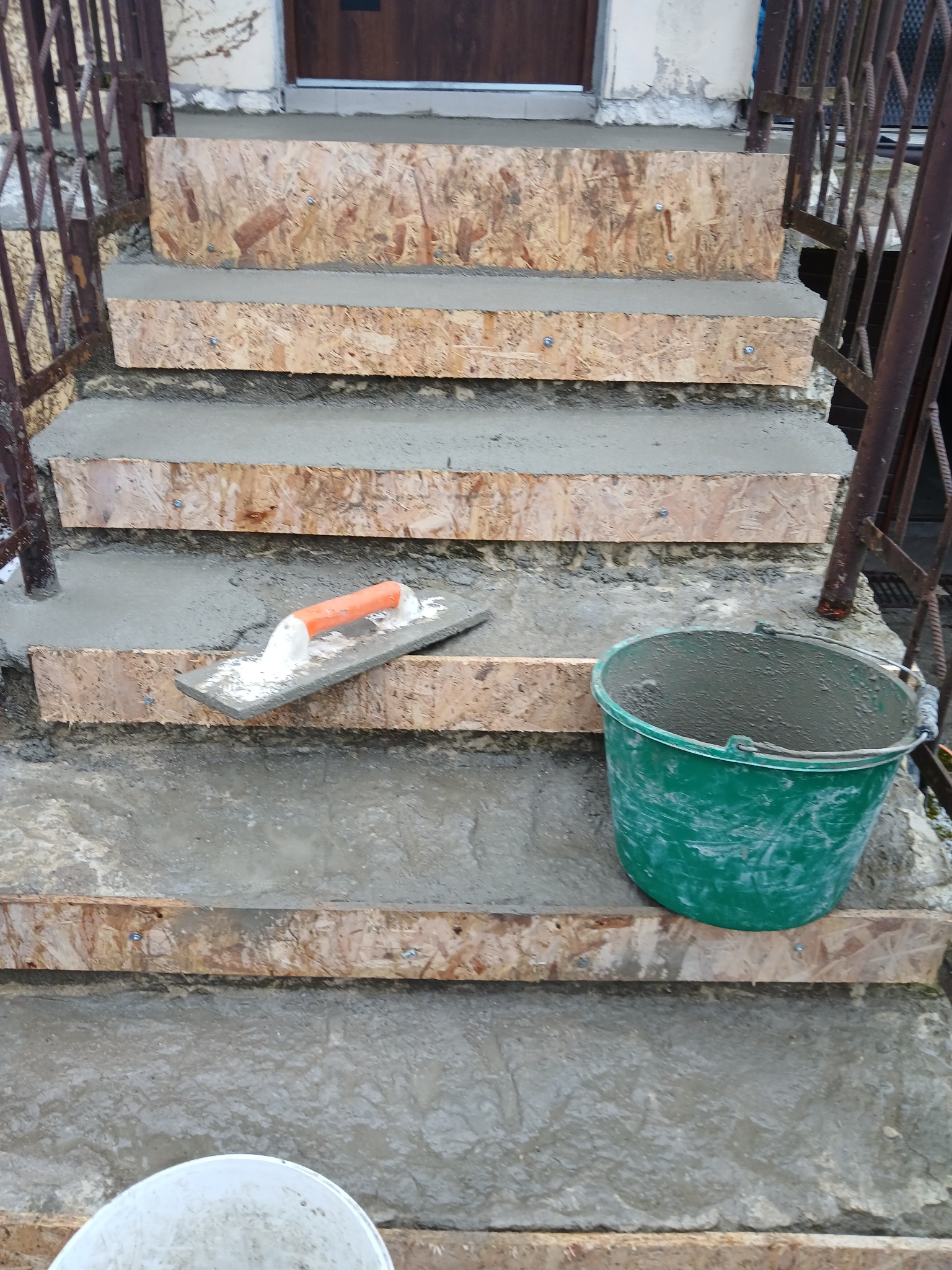 Schody wejściowe w trakcie betonowania, z deskowaniem z płyty OSB. Widoczne narzędzia: kielnia z zaprawą i zielone wiadro. Metalowa barierka po bokach.