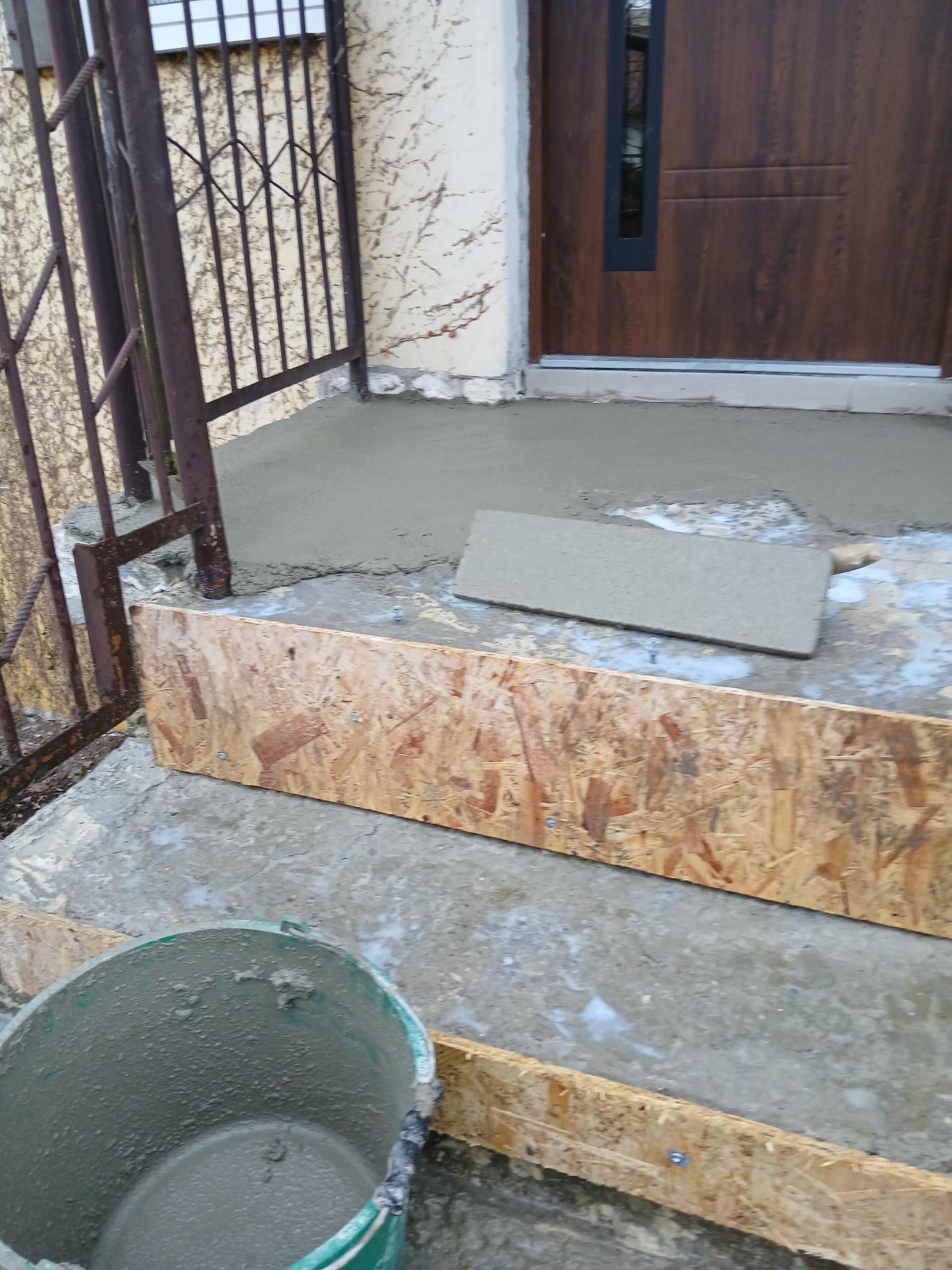 Wylewka betonowa przed wejściem do domu, szalunek z desek, wiadro z zaprawą. Widoczna brama wejściowa i elewacja budynku z roślinnością.