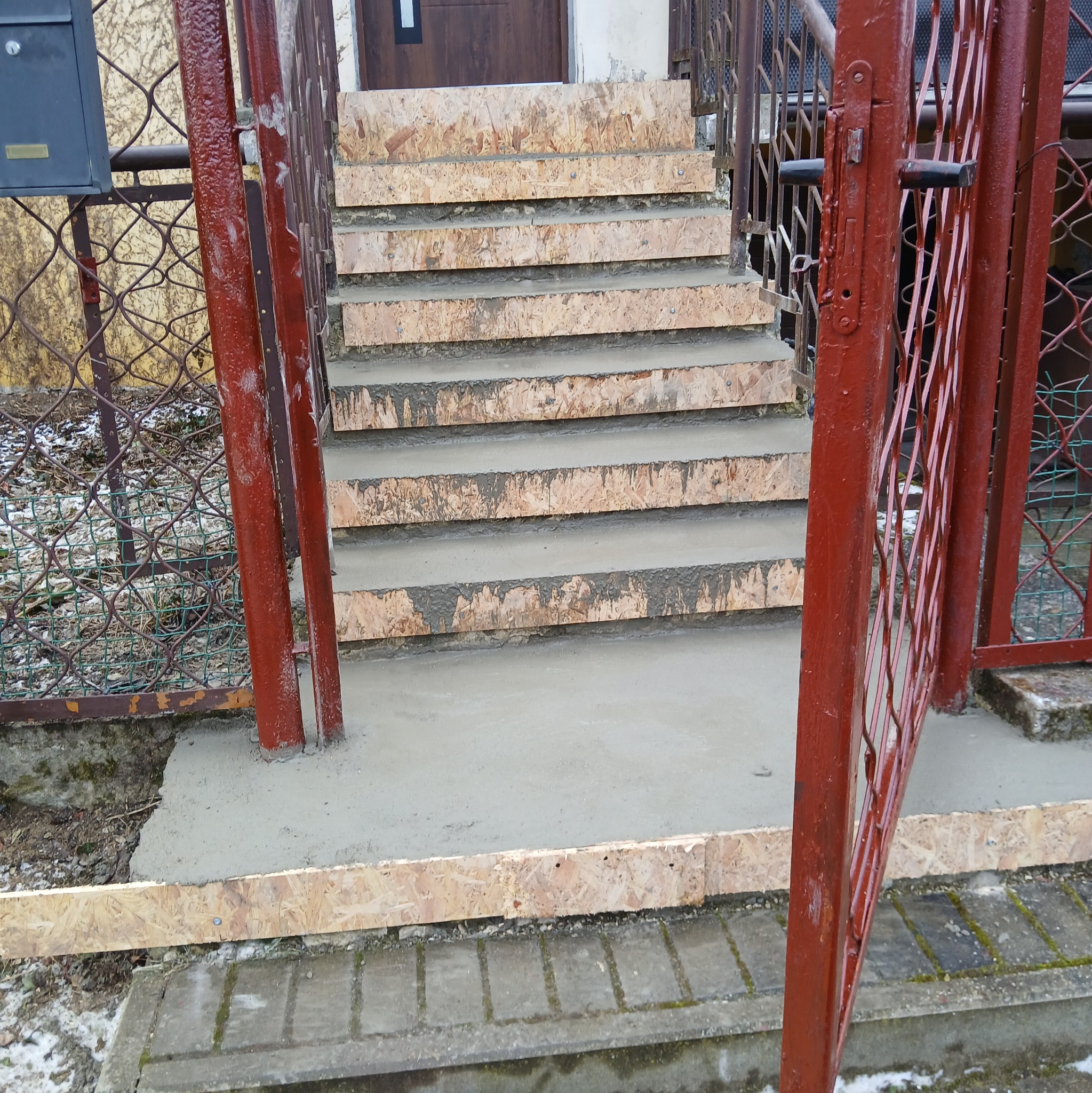 Betonowe schody w trakcie budowy z widocznymi deskami szalunkowymi, czerwona metalowa furtka i siatka ogrodzeniowa w tle.
