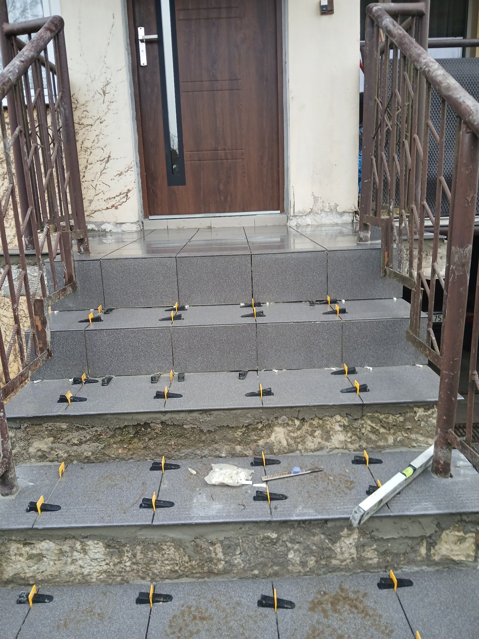 Montaż płytek na schodach wejściowych z użyciem klipsów poziomujących. Widoczne narzędzia: poziomica, nóż. Częściowo wykończone schody z szarych płytek.