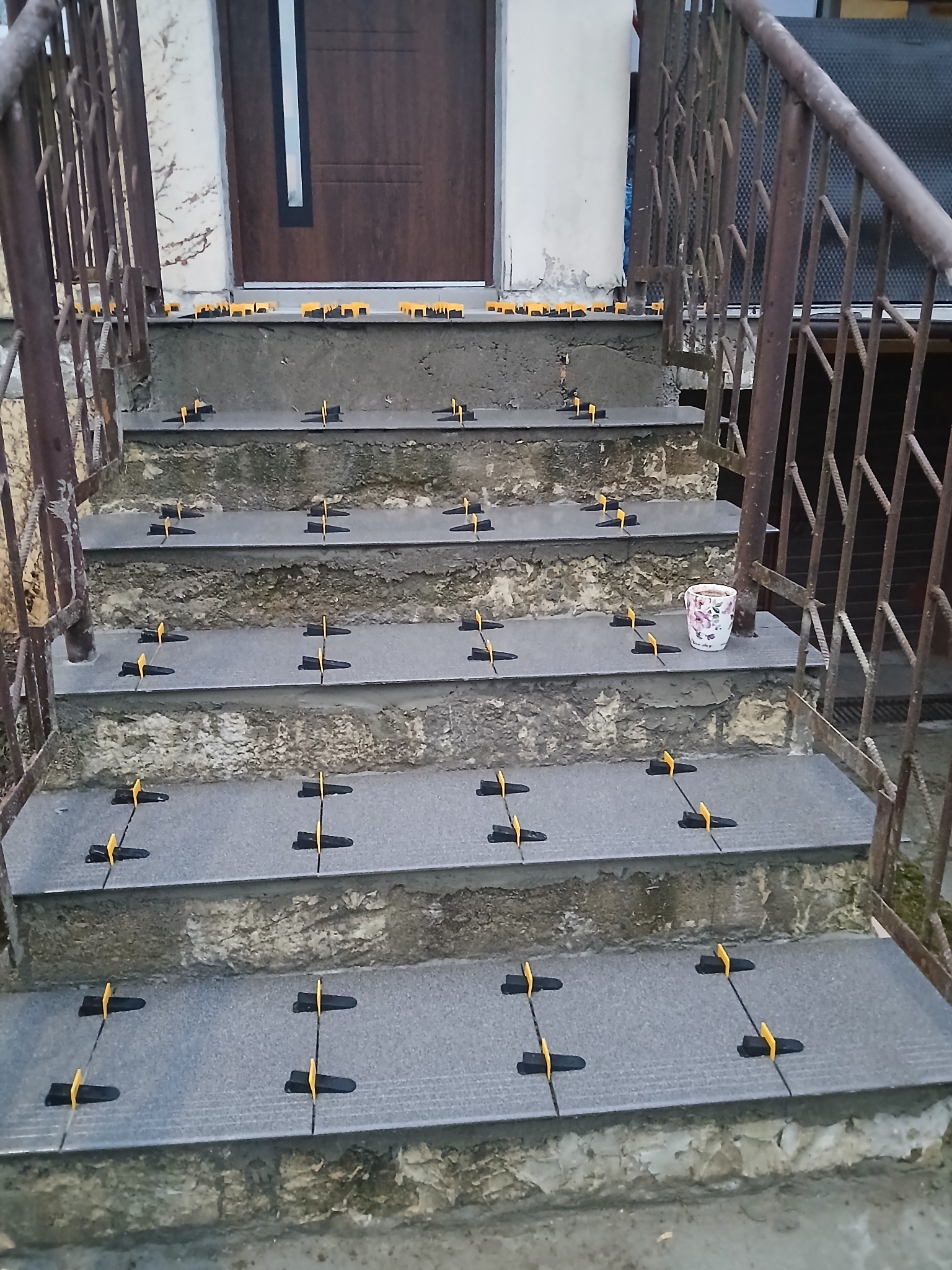 Montaż płytek na schodach wejściowych z użyciem klipsów poziomujących. Widoczne metalowe barierki i drzwi wejściowe. Kubek z kwiatami na jednym ze stopni.