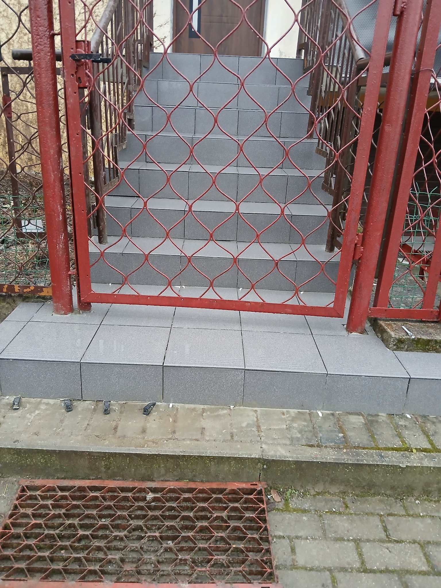 Czerwona, metalowa furtka z siatki na tle schodów wyłożonych szarą kostką. Widoczny fragment kratki ściekowej i płytek na schodach.