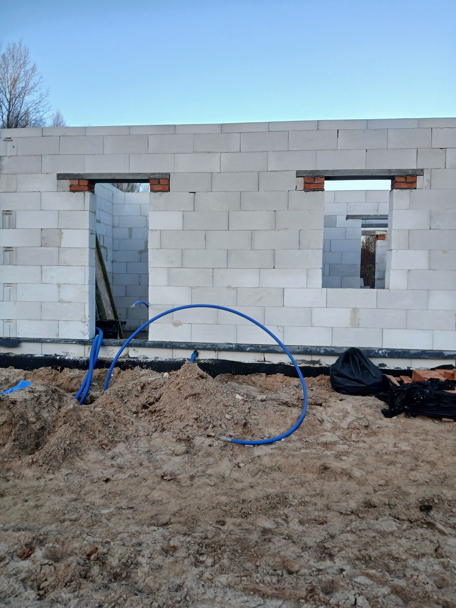 Wznoszenie ściany z betonu komórkowego z otworami okiennymi i drzwiowymi. Widoczne niebieskie rury instalacyjne i czarna folia izolacyjna u podstawy ściany.