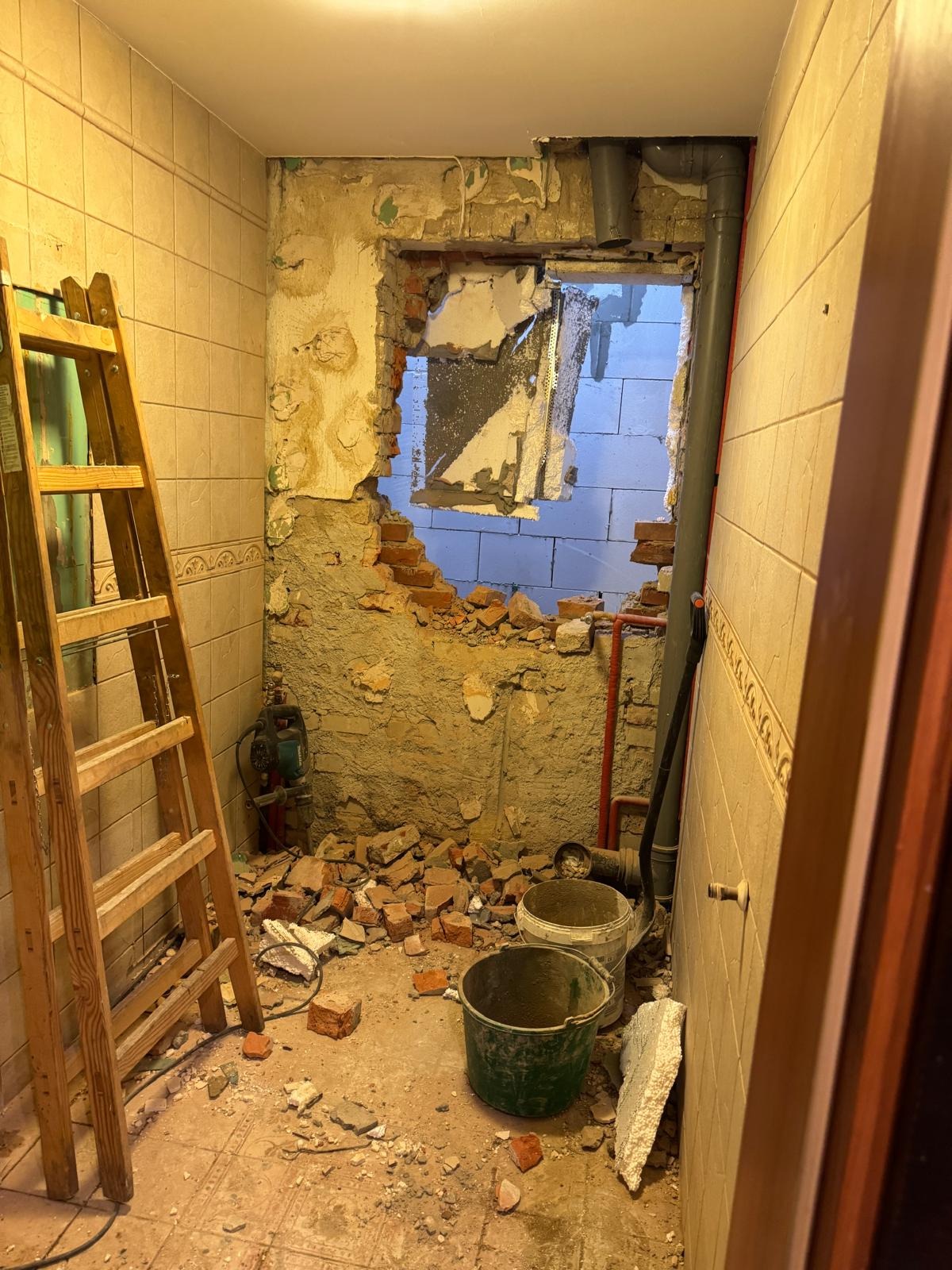 Wnętrze łazienki w trakcie remontu: usunięte płytki, gruz, drabina oparta o ścianę, widoczne instalacje. Otwór w ścianie z widokiem na zewnątrz.
