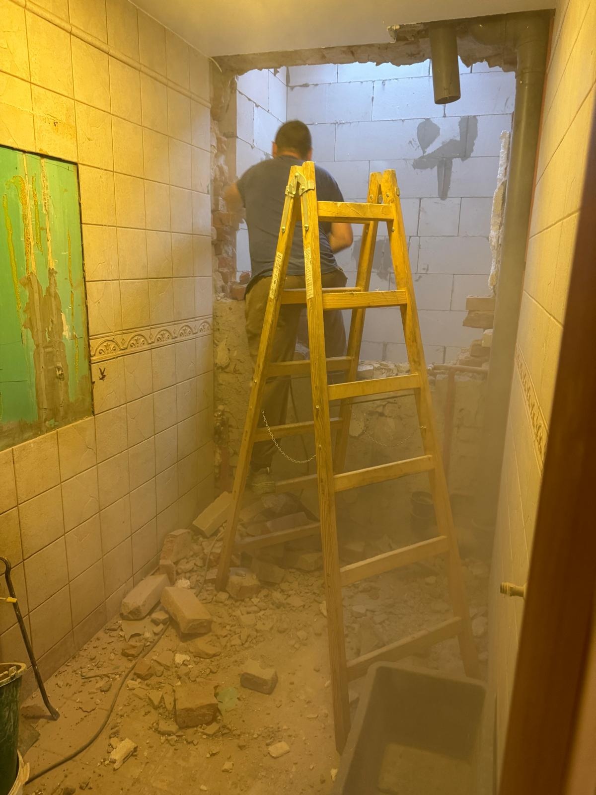 Pracownik na drabinie wyburza ścianę z pustaków w łazience z beżowymi płytkami, widoczne gruz i instalacje. Prace remontowe w trakcie realizacji.