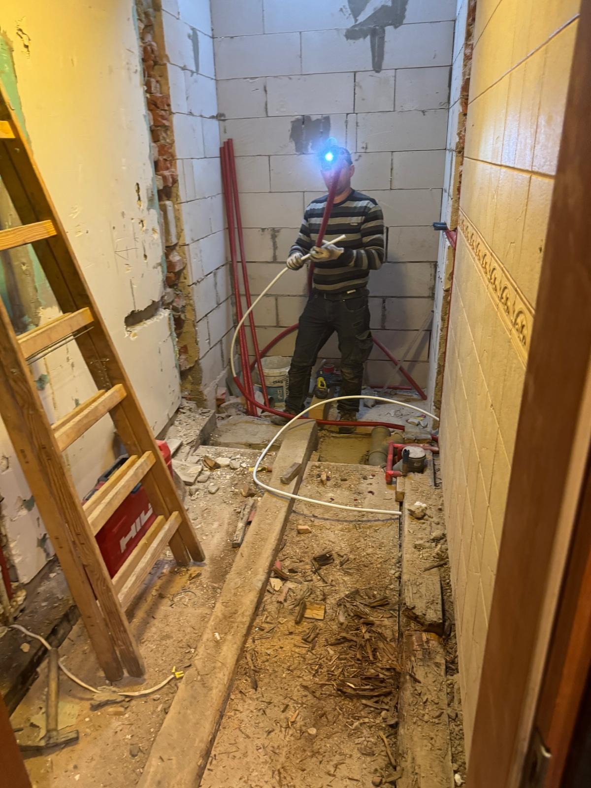 Instalator z czołówką montuje rury w surowym wnętrzu łazienki z widoczną drabiną i fragmentami ścian z pustaków oraz płytek. Etap prac instalacyjnych.