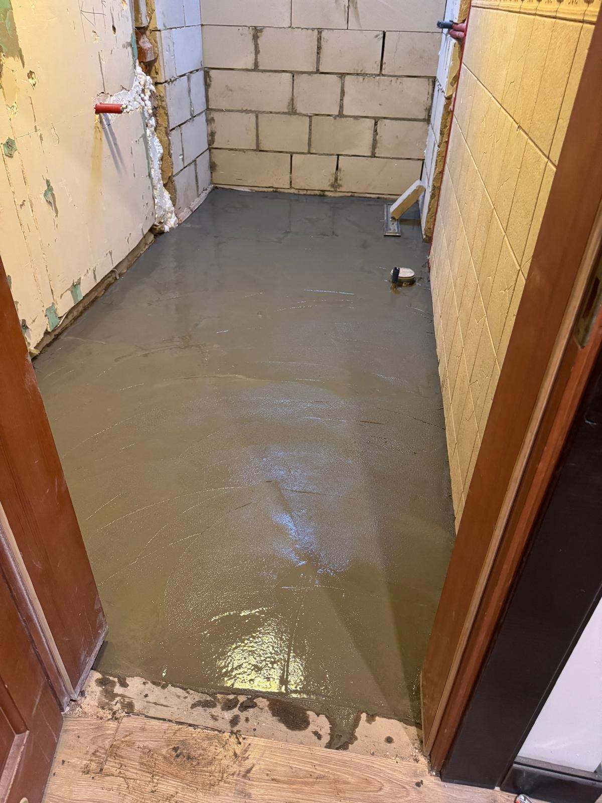 Wylany, świeży podkład podłogowy w pomieszczeniu z fragmentami ścian z bloczków i płytek. Widoczne narzędzia do rozprowadzania wylewki.