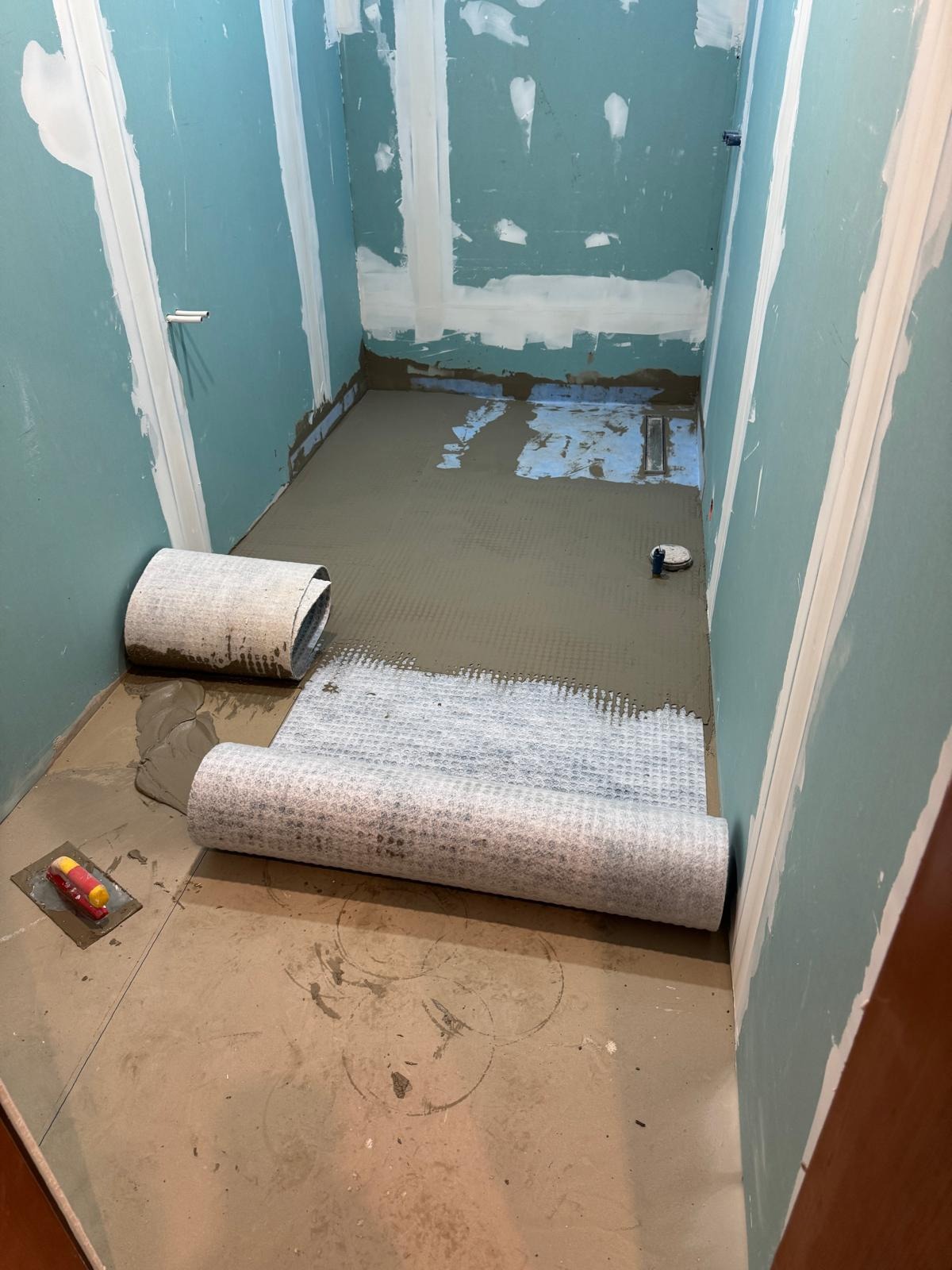 Przygotowanie podłoża pod płytki w łazience: rozłożona mata izolacyjna na warstwie wylewki, widoczne narzędzia i surowe ściany z płyt gipsowych.