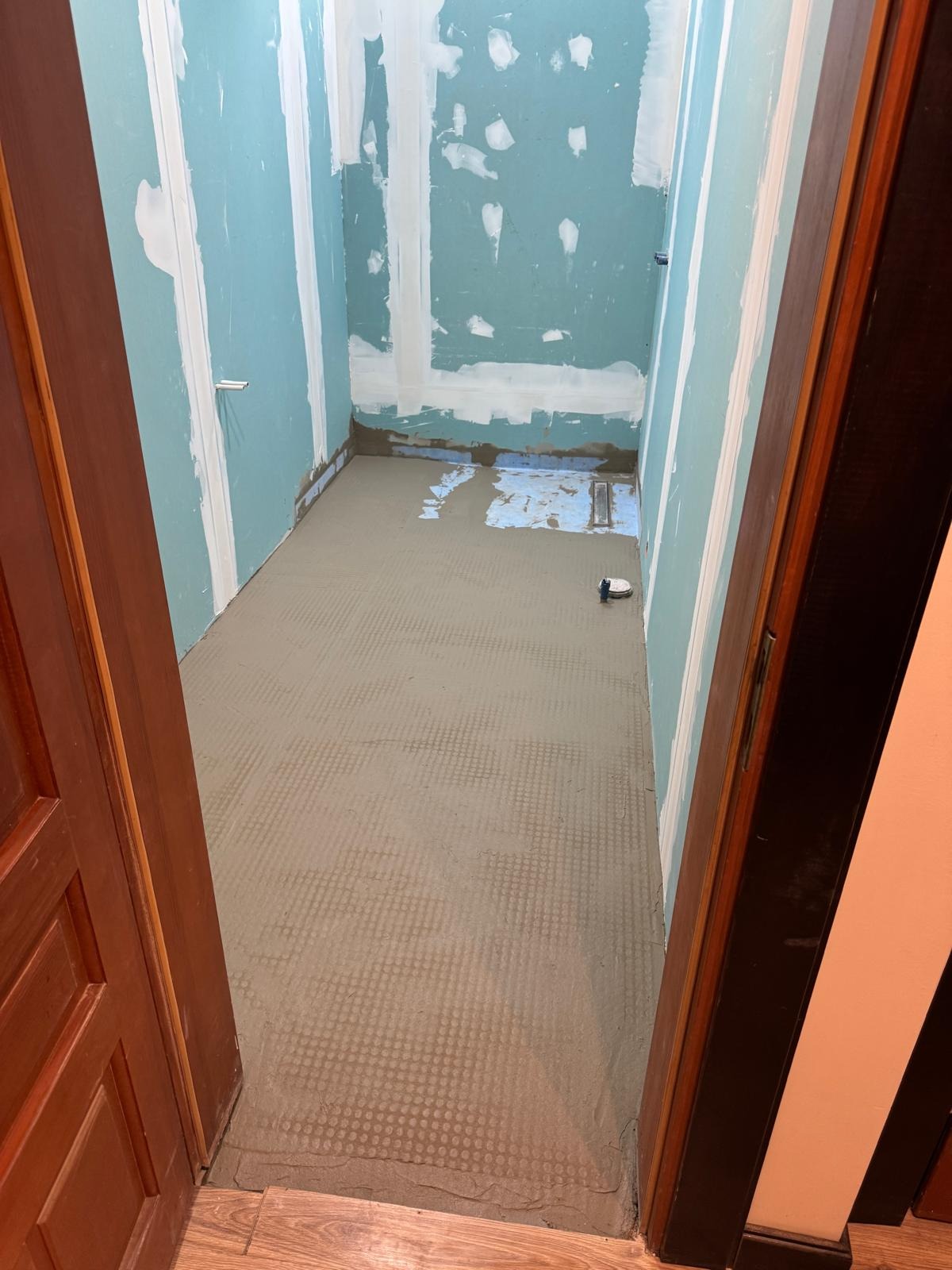 Przygotowanie podłoża w łazience: wylewka samopoziomująca na siatce, ściany z płyt gipsowo-kartonowych, widoczny odpływ liniowy. Drzwi wejściowe z prawej strony kadru.