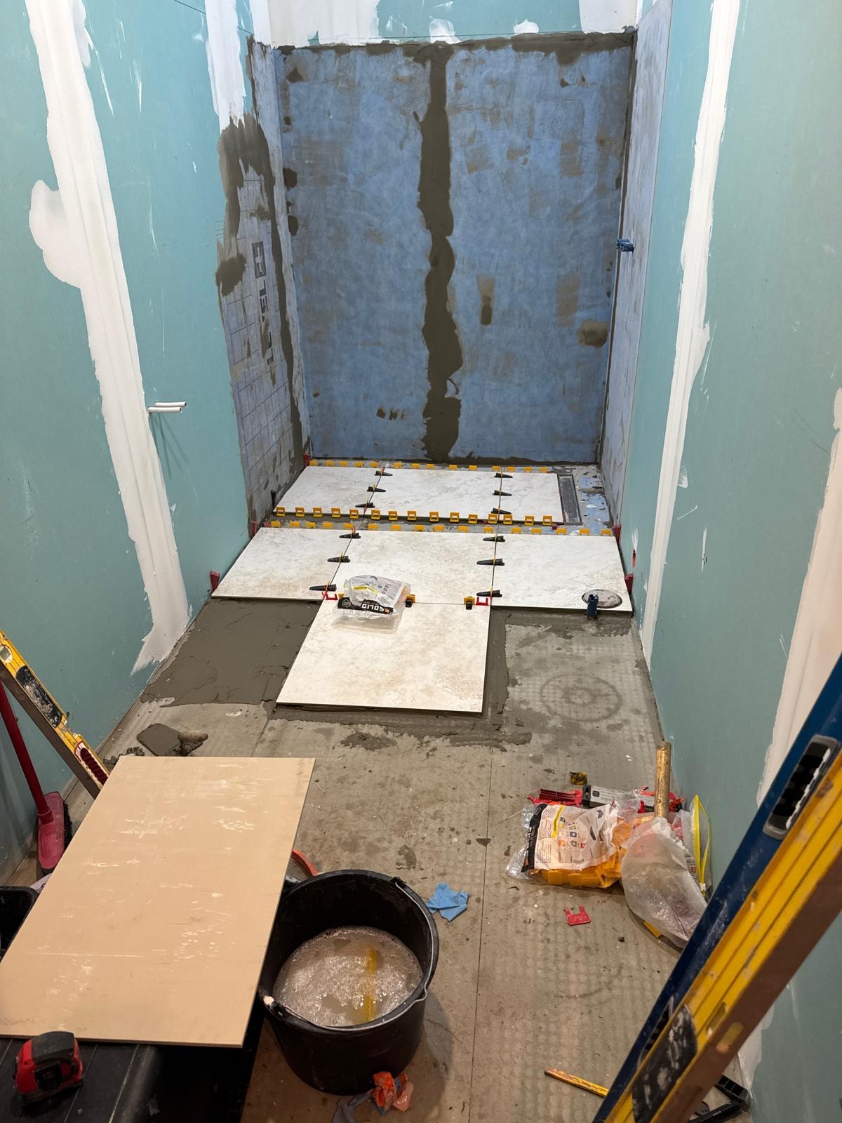 Wnętrze łazienki w trakcie remontu: częściowo ułożone płytki podłogowe z systemem poziomowania, przygotowane ściany, widoczne narzędzia i materiały budowlane.