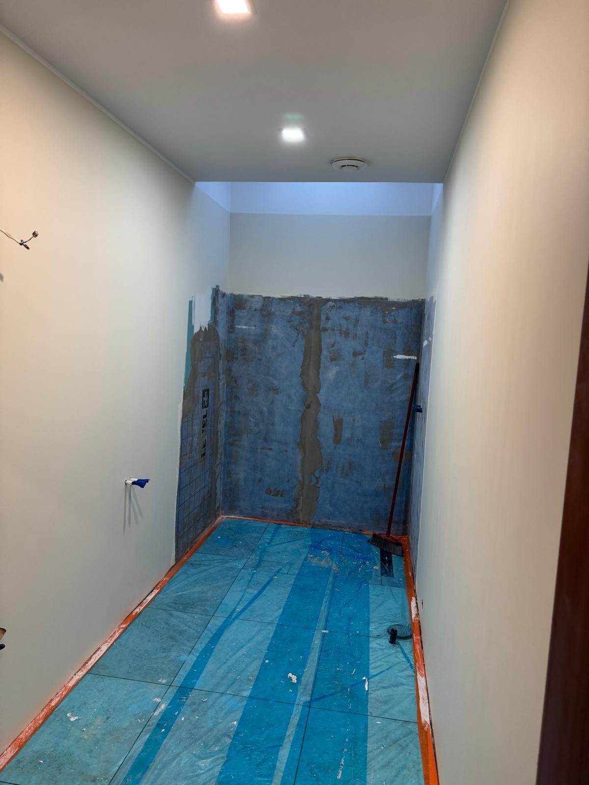 Wnętrze w trakcie remontu: zabezpieczona podłoga, przygotowane ściany pod płytki, widoczna instalacja. Prace wykończeniowe w łazience lub innym pomieszczeniu.