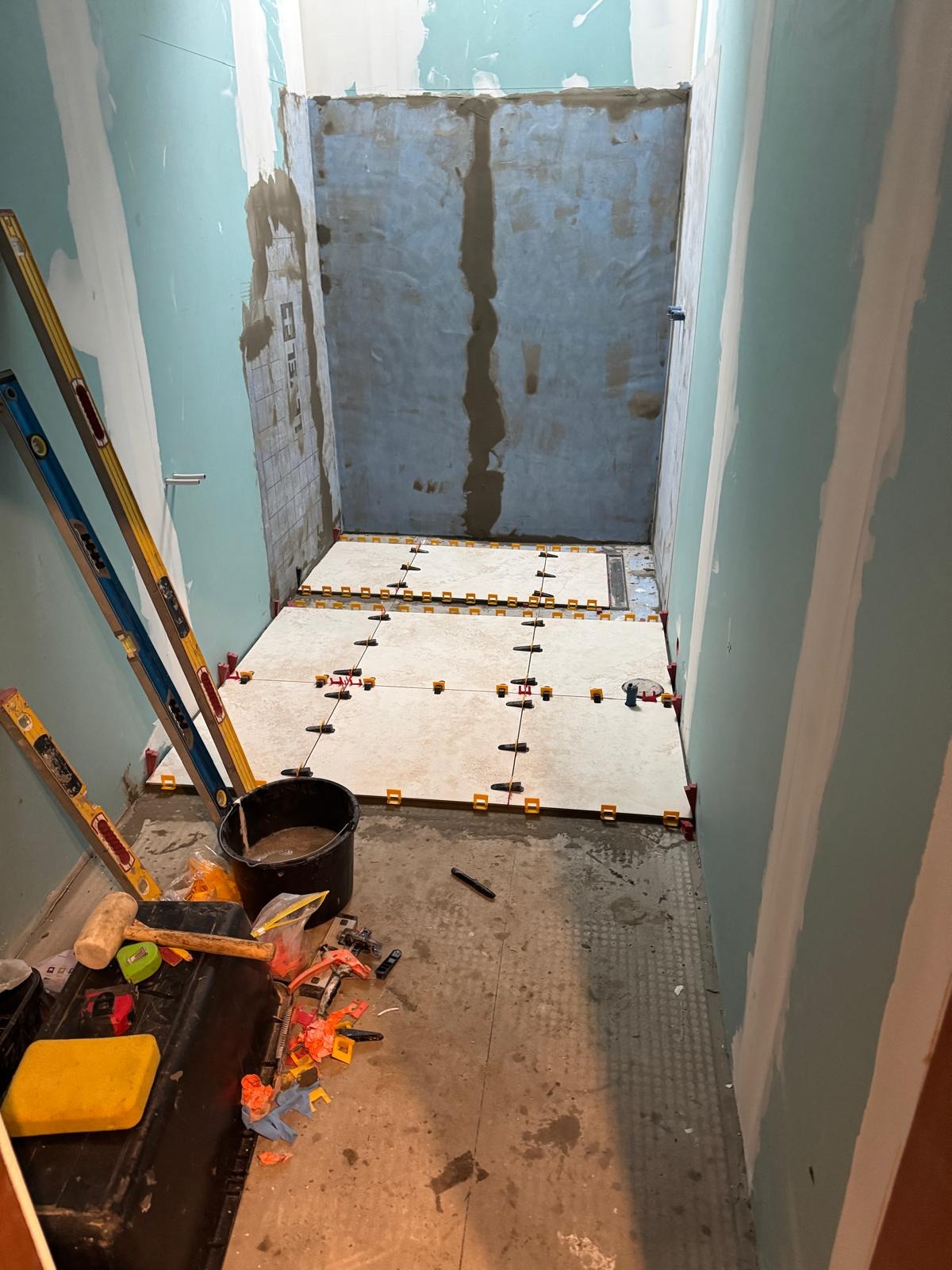 Wnętrze łazienki w trakcie remontu: ułożone płytki podłogowe z systemem poziomowania, widoczne narzędzia i poziomice. Ściany przygotowane do dalszych prac wykończeniowych.