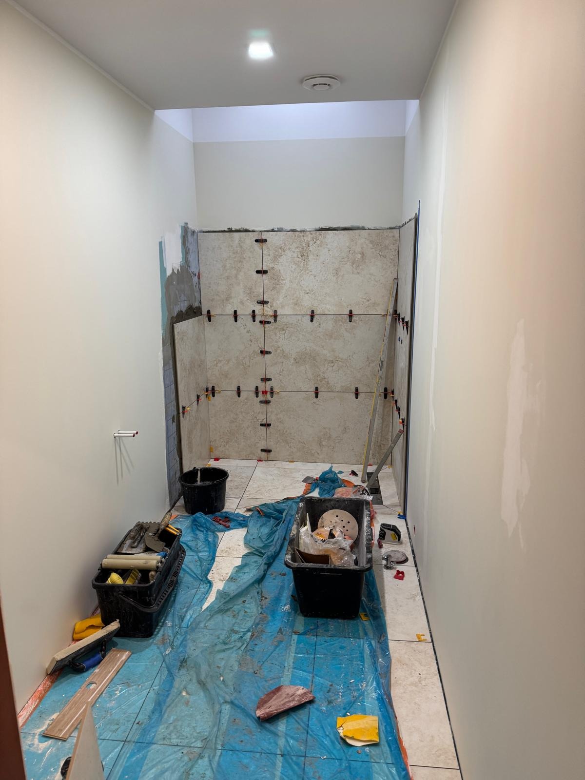 Wnętrze łazienki w trakcie remontu: ściany wyłożone płytkami z użyciem krzyżyków dystansowych, narzędzia i akcesoria na podłodze zabezpieczonej folią.