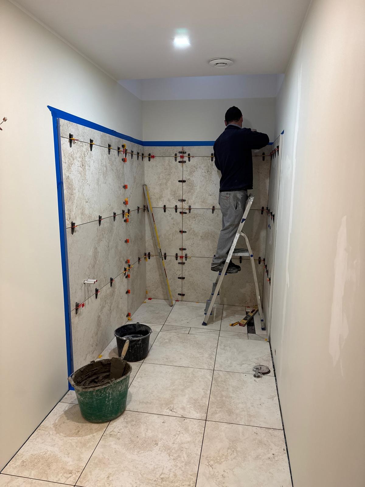 Układanie beżowych płytek w łazience z użyciem poziomicy i systemu poziomowania. Mężczyzna na drabinie montuje płytki. Widoczne wiaderka z zaprawą.