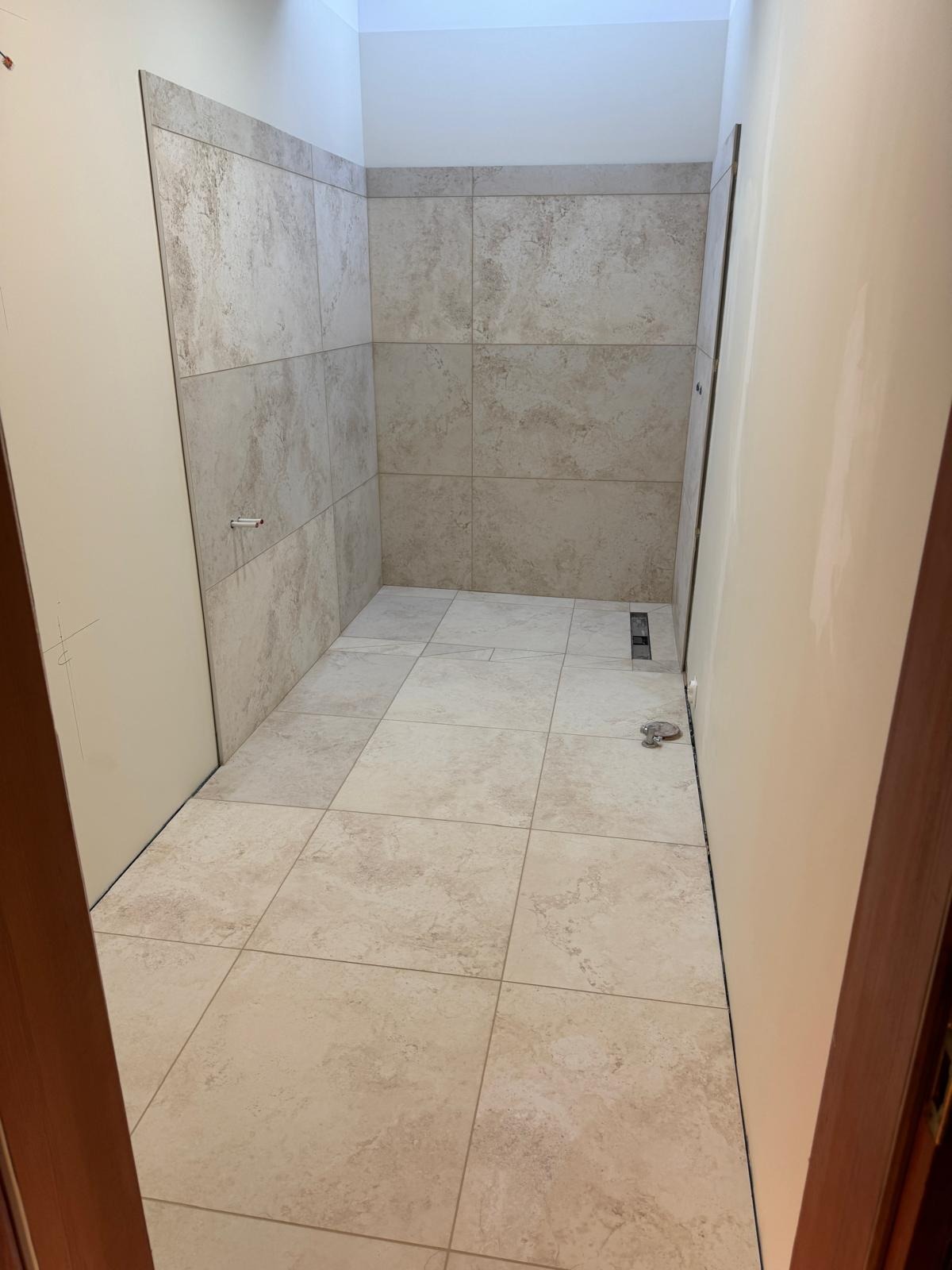 Wykończona łazienka z beżowymi płytkami na ścianach i podłodze. Widoczny odpływ liniowy i fragment drzwi. Neutralna kolorystyka i minimalistyczny design.