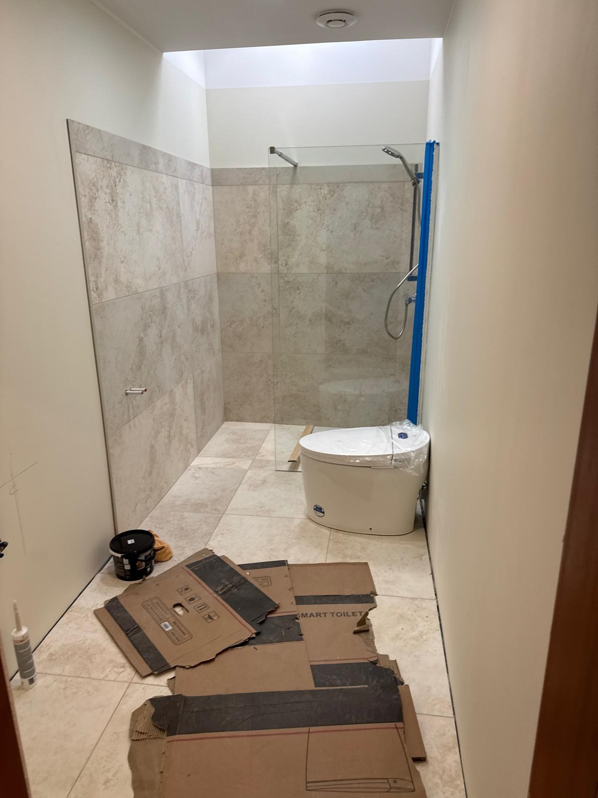 Wnętrze łazienki w trakcie wykańczania: ułożone płytki, zamontowana kabina prysznicowa z niebieskim zabezpieczeniem, toaleta w folii, kartony na podłodze, widok z góry.