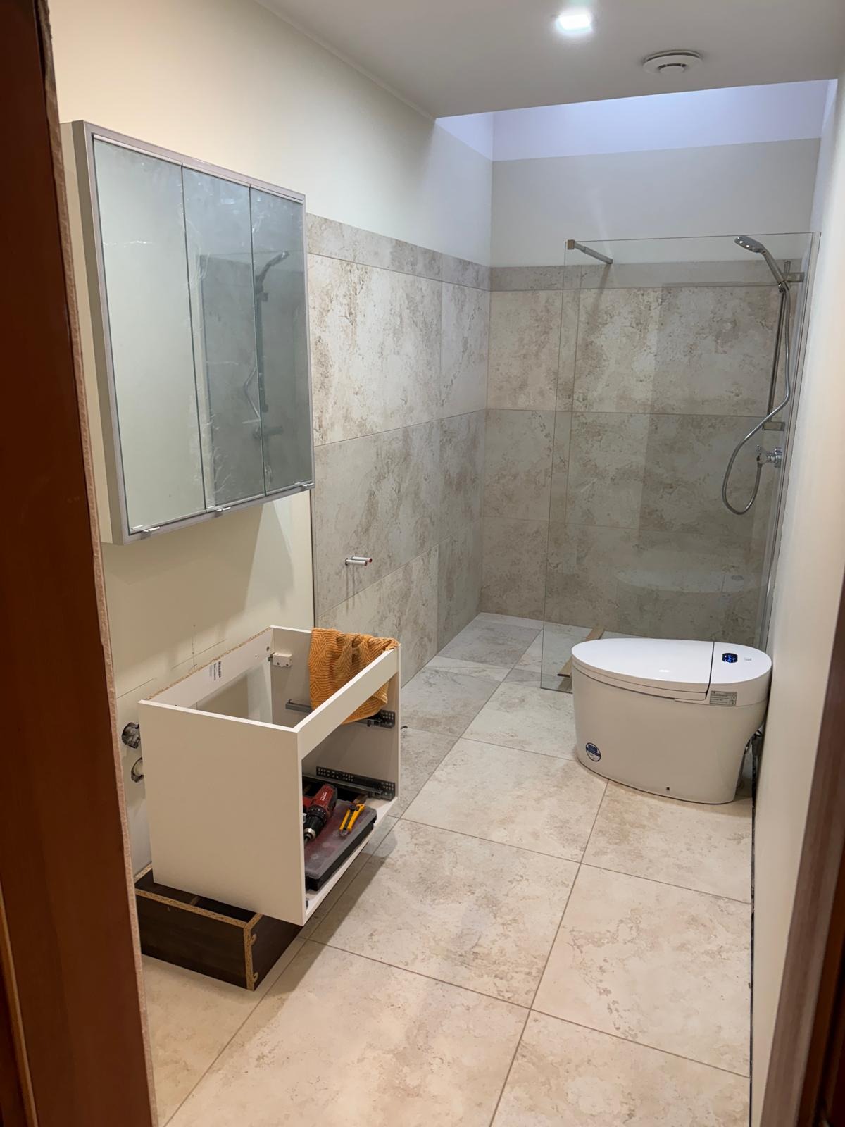 Remont łazienki: montaż szafki pod umywalkę, widoczna kabina prysznicowa i toaleta. Na szafce leżą narzędzia. Beżowe płytki na podłodze i ścianach.