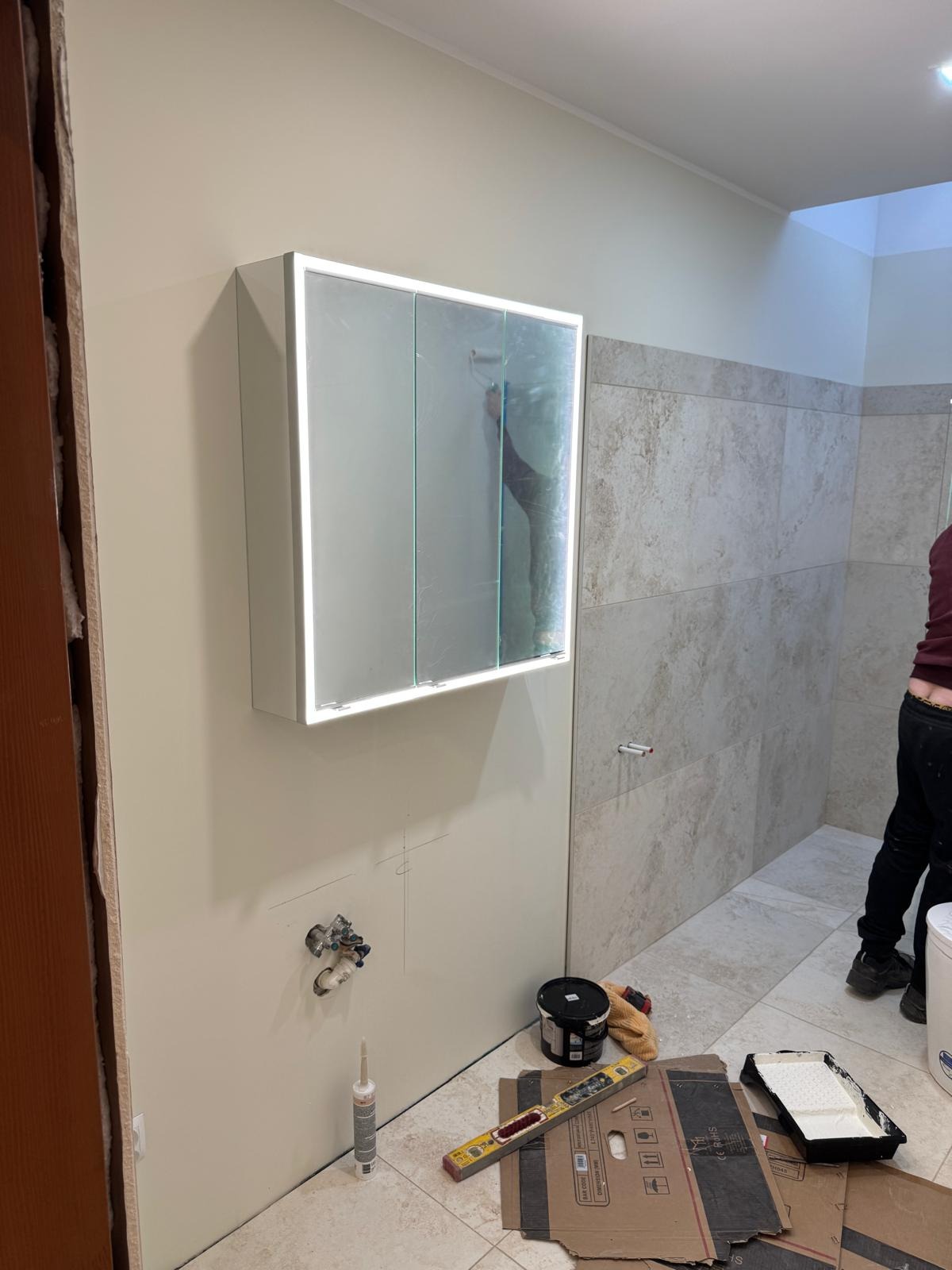 Montaż szafki z lustrem i oświetleniem LED w łazience z beżowymi płytkami. Widoczne narzędzia i materiały budowlane. Częściowo widoczna osoba wykonująca prace.