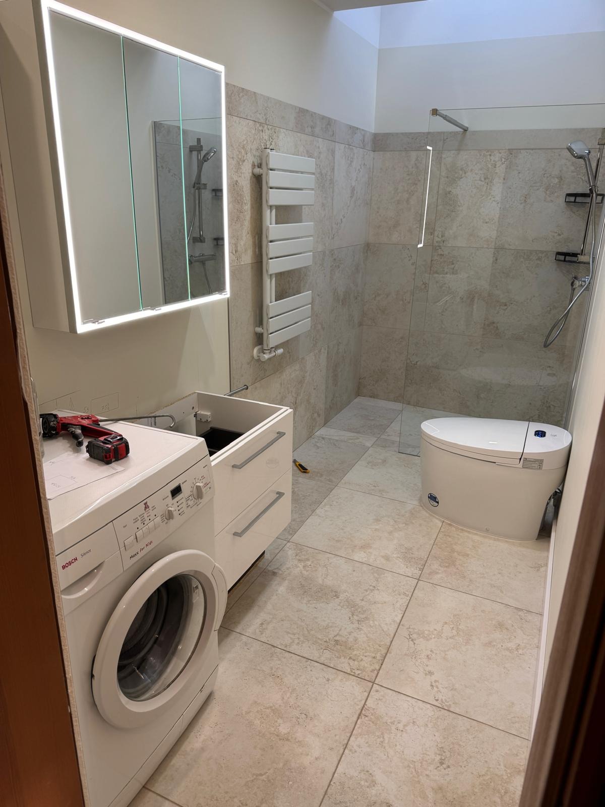 Nowoczesna łazienka w trakcie montażu: biała pralka Bosch obok szafki z otwartą szufladą, narzędzia na pralce, kabina prysznicowa w tle, beżowe płytki podłogowe i ścienne.
