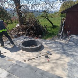 S&S - Budowa paleniska z kostki brukowej na posesji. Widoczne narzędzia i ułożona kostka. W tle drzewo i sterta gałęzi, co dodaje naturalnego uroku.