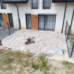 S&S - Nowy taras z kostki brukowej w odcieniach beżu i szarości, częściowo otoczony metalowym ogrodzeniem. Na kostce stoi zagęszczarka, a w tle widoczny fragment domu.