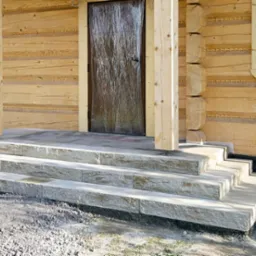 Kamienne schody wejściowe do drewnianego domu z bali. Trzy stopnie prowadzą do drzwi. Widoczne detale drewnianej konstrukcji i otoczenie domu.