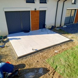 S&S - Jasne, kwadratowe płyty tarasowe ułożone przy budynku. Widoczny fragment pracownika przygotowującego podłoże, narzędzia i fragment elewacji z roletami.