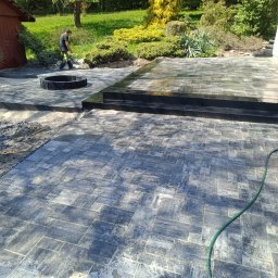 S&S - Układanie kostki brukowej w ogrodzie z czarnym obramowaniem paleniska. Pracownik w tle. Częściowo mokra nawierzchnia. Węże ogrodowe na kostce brukowej.