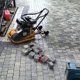 S&S - Proces układania szarej kostki brukowej na podjeździe. Widoczna zagęszczarka, narzędzia i częściowo ułożona nawierzchnia. Prace w toku.