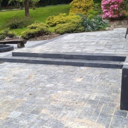 S&S - Ułożona kostka brukowa w ogrodzie z dwoma stopniami, paleniskiem i drewnianą ławką. W tle budynek z kamienia i roślinność.