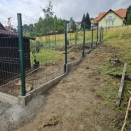 Montaż ogrodzenia panelowego z podmurówką betonową na nierównym terenie. Widoczne słupki w kolorze ciemnej zieleni i siatka w kolorze grafitowym, dom w tle.