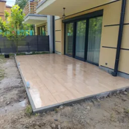 Nowoczesny taras z płytek imitujących drewno, wykończony szarą obwódką, przylegający do budynku z dużymi przesuwnymi drzwiami i balkonem na piętrze.