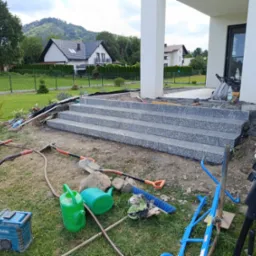 Granitowe schody wejściowe do nowoczesnego domu w trakcie montażu. W tle widoczne domy jednorodzinne i zieleń. Narzędzia i materiały porozrzucane wokół.