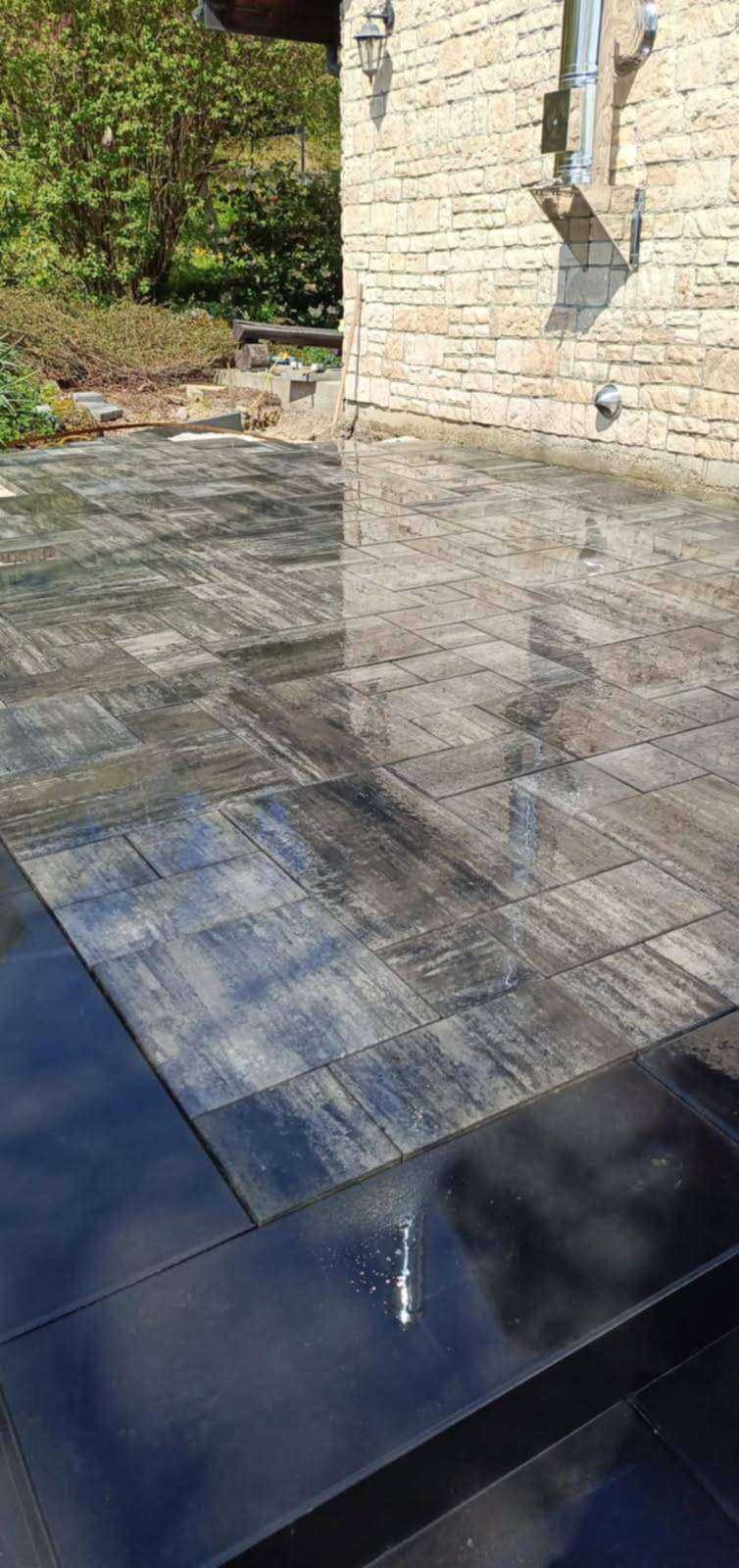 Świeżo ułożona, mokra kostka brukowa w odcieniach szarości i czerni, odbijająca światło, przylegająca do kamiennej ściany budynku i zieleni w tle.