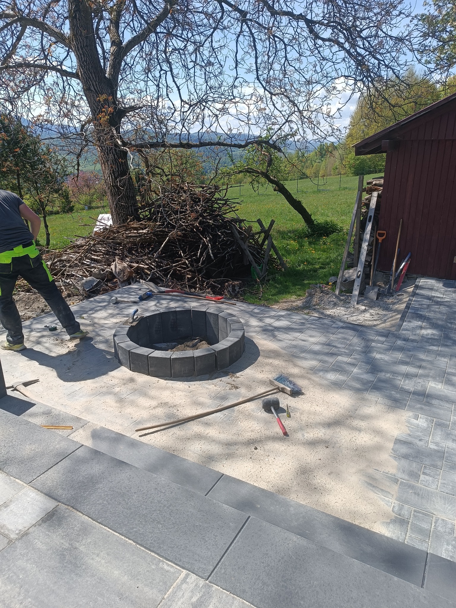 Budowa paleniska z kostki brukowej na posesji. Widoczne narzędzia i ułożona kostka. W tle drzewo i sterta gałęzi, co dodaje naturalnego uroku.