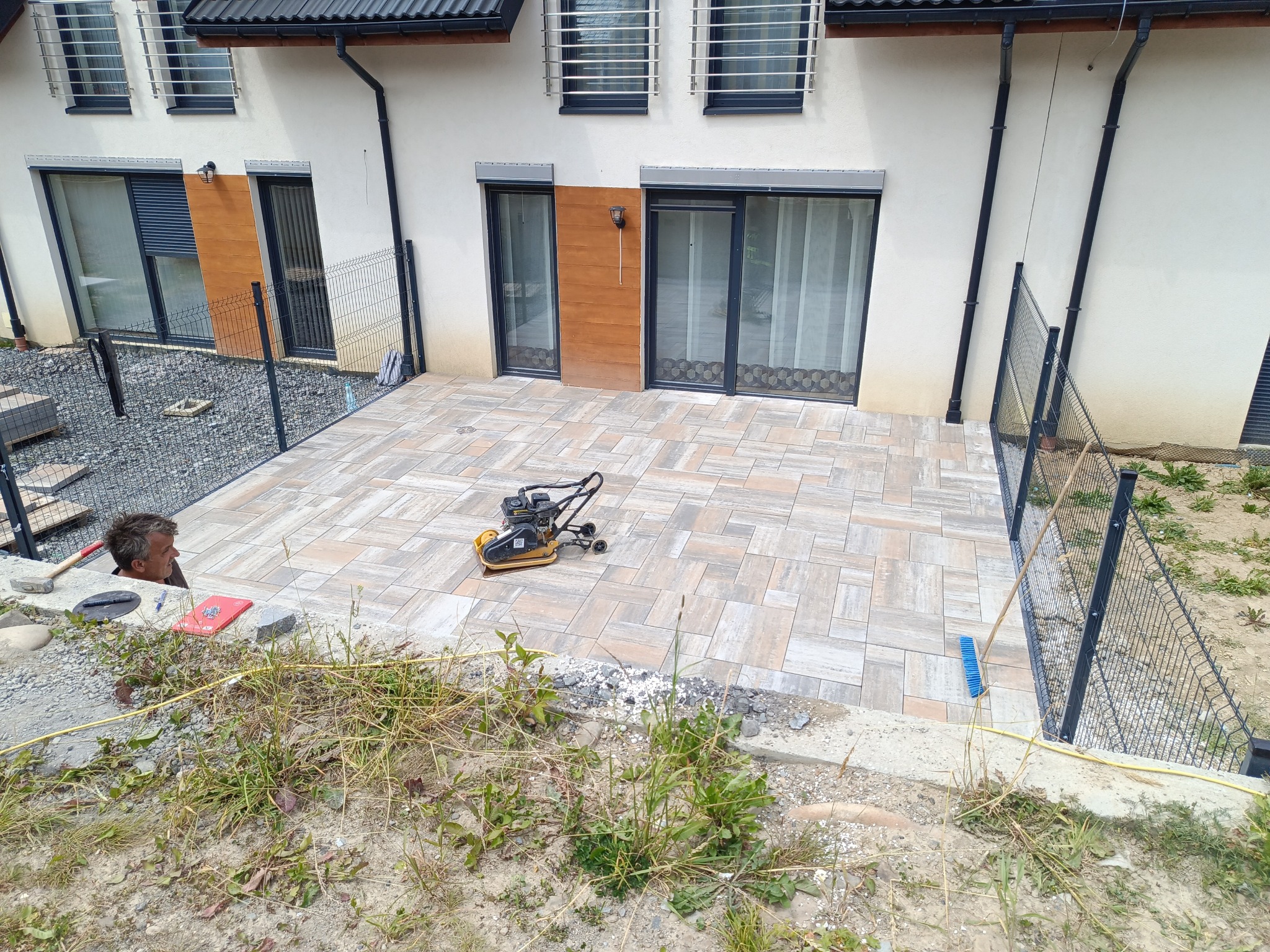 Nowy taras z kostki brukowej w odcieniach beżu i szarości, częściowo otoczony metalowym ogrodzeniem. Na kostce stoi zagęszczarka, a w tle widoczny fragment domu.
