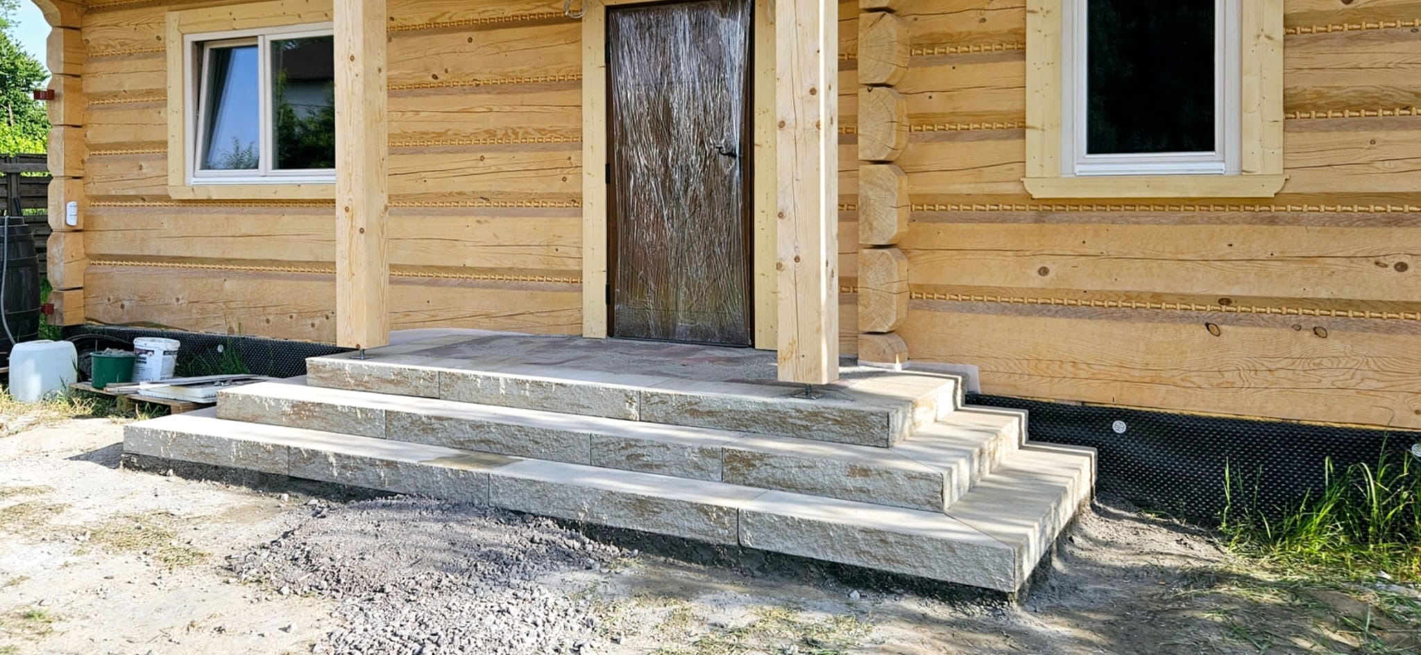 Kamienne schody wejściowe do drewnianego domu z bali. Trzy stopnie prowadzą do drzwi. Widoczne detale drewnianej konstrukcji i otoczenie domu.