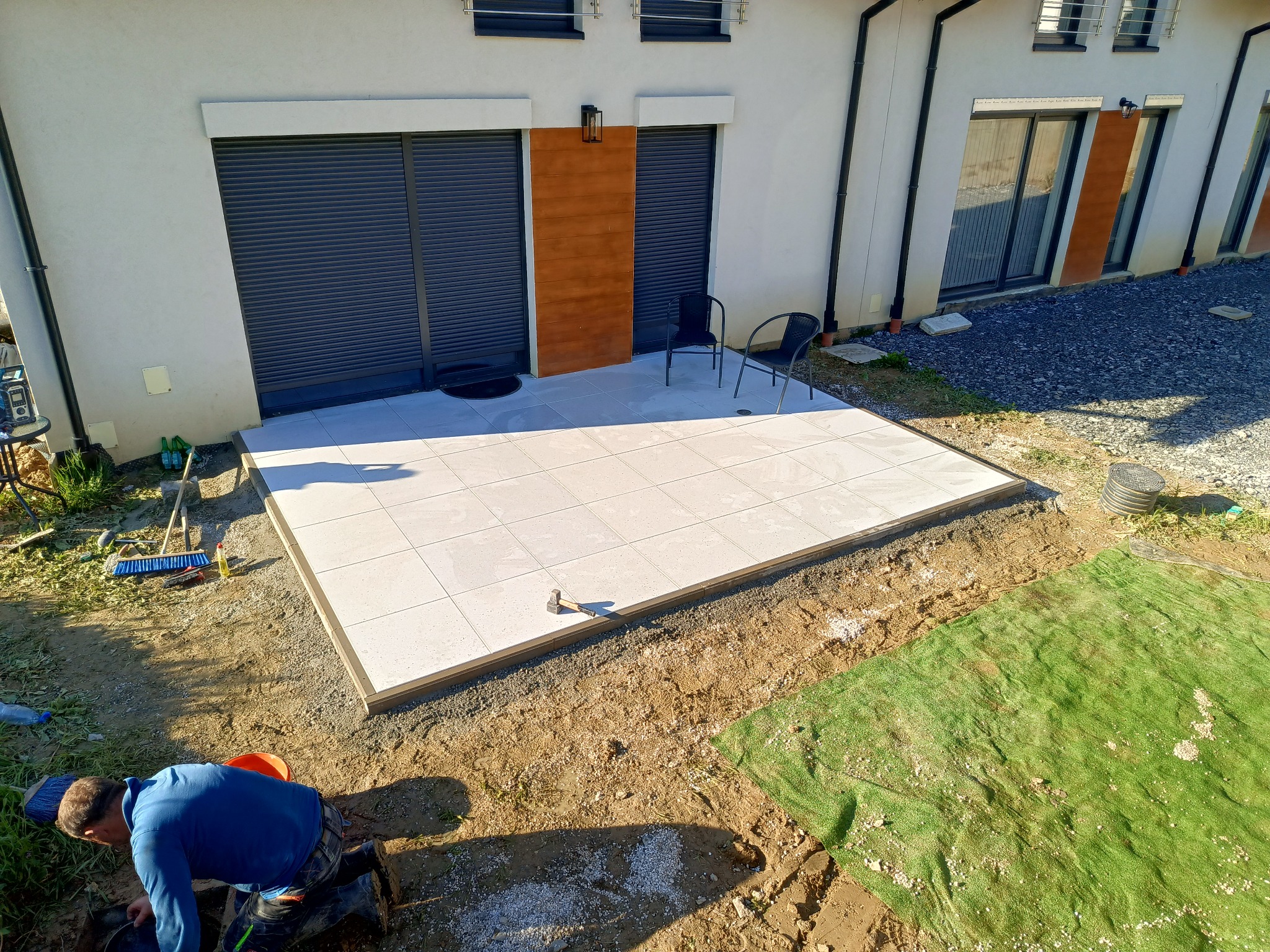 Jasne, kwadratowe płyty tarasowe ułożone przy budynku. Widoczny fragment pracownika przygotowującego podłoże, narzędzia i fragment elewacji z roletami.