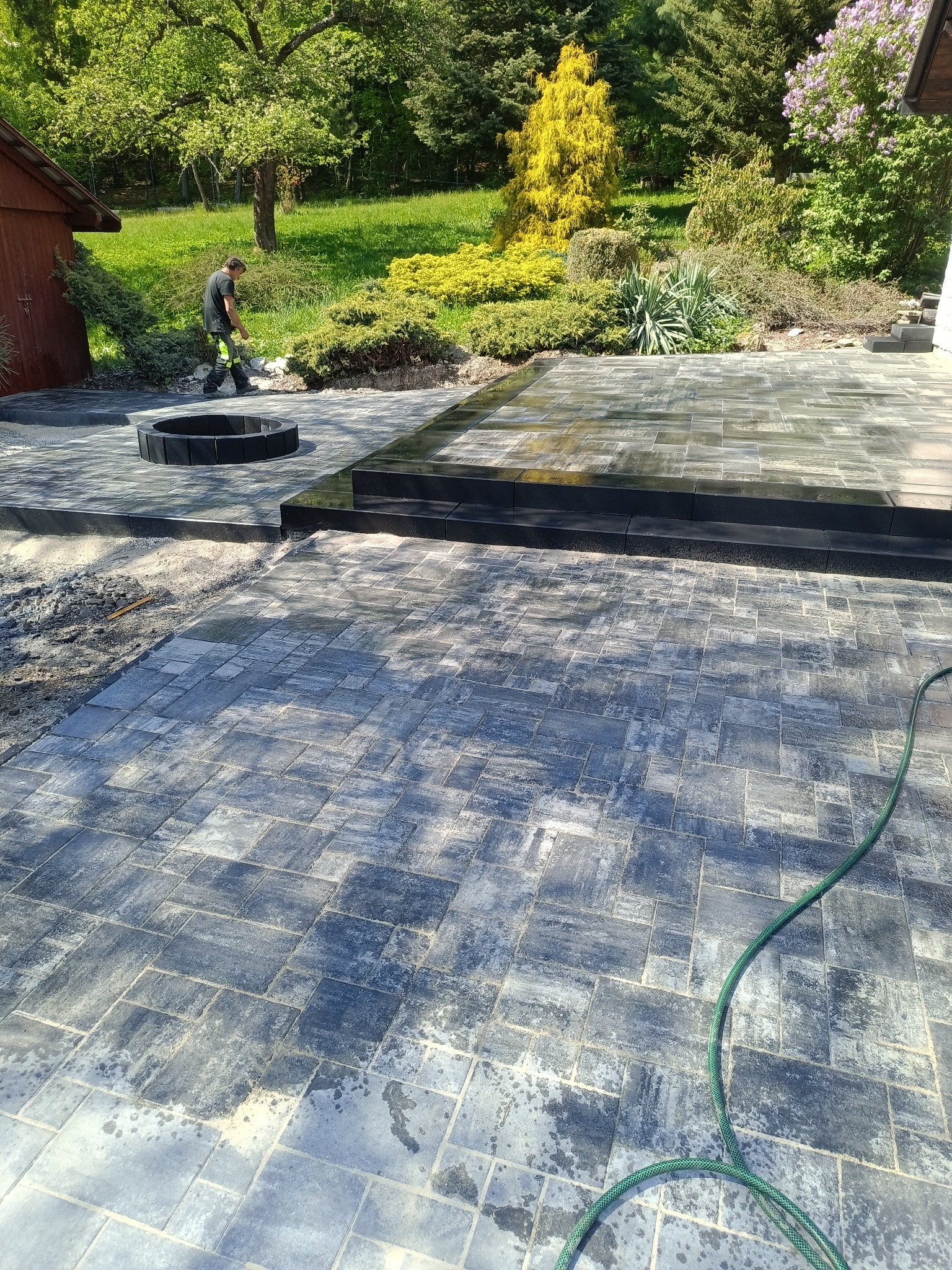 Układanie kostki brukowej w ogrodzie z czarnym obramowaniem paleniska. Pracownik w tle. Częściowo mokra nawierzchnia. Węże ogrodowe na kostce brukowej.