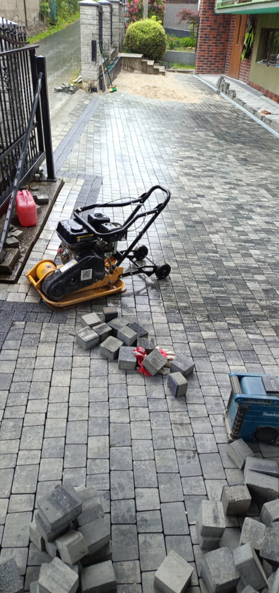Proces układania szarej kostki brukowej na podjeździe. Widoczna zagęszczarka, narzędzia i częściowo ułożona nawierzchnia. Prace w toku.