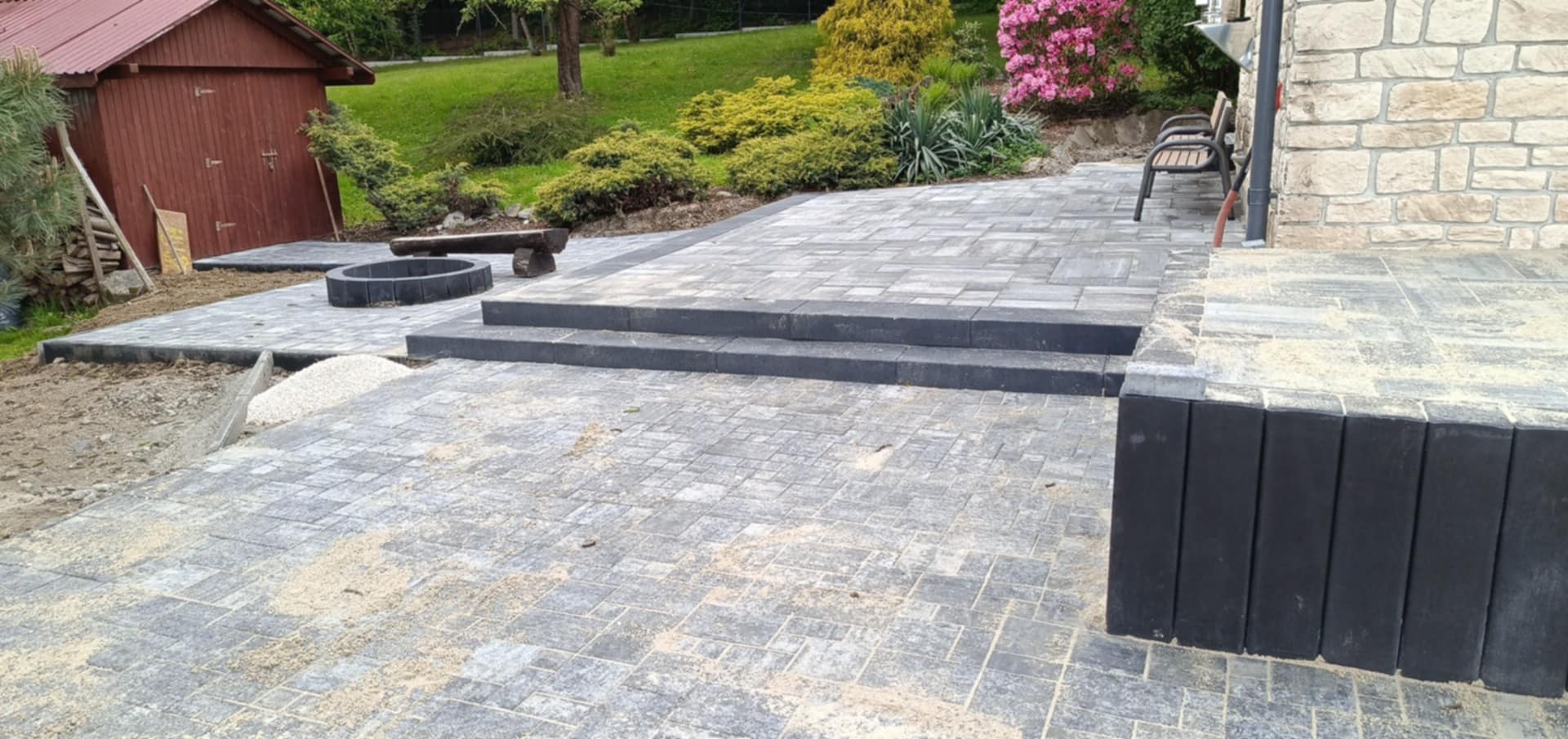 Ułożona kostka brukowa w ogrodzie z dwoma stopniami, paleniskiem i drewnianą ławką. W tle budynek z kamienia i roślinność.