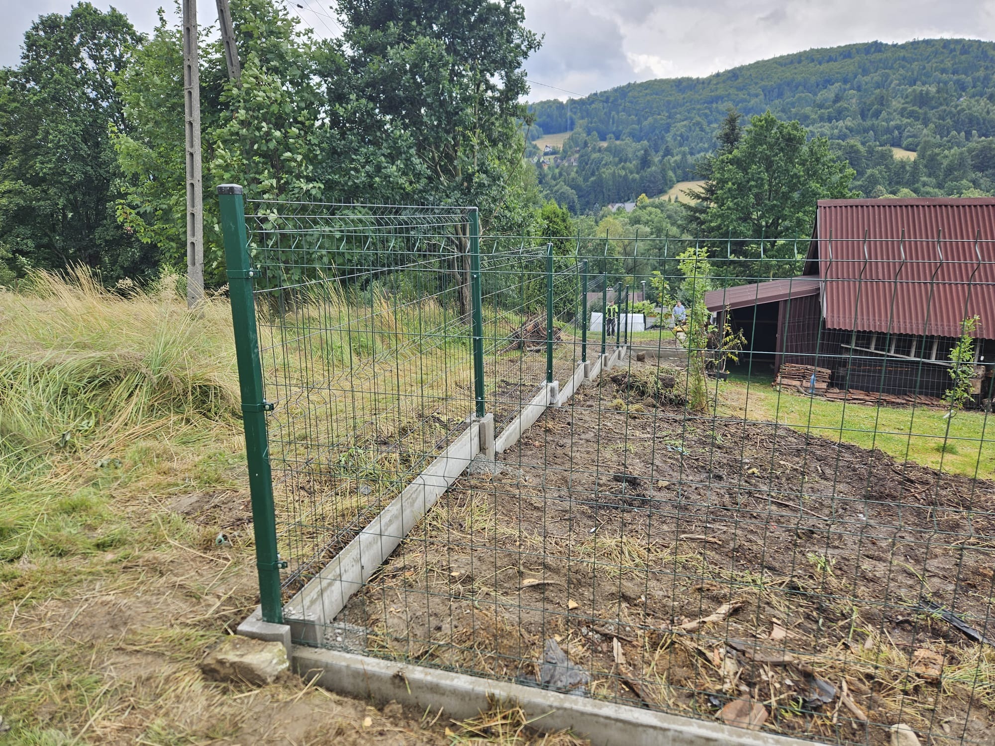 Montaż ogrodzenia z siatki na betonowej podmurówce, widok na pagórkowaty krajobraz. Słupki metalowe, zielone. Prace w toku, teren przygotowany.